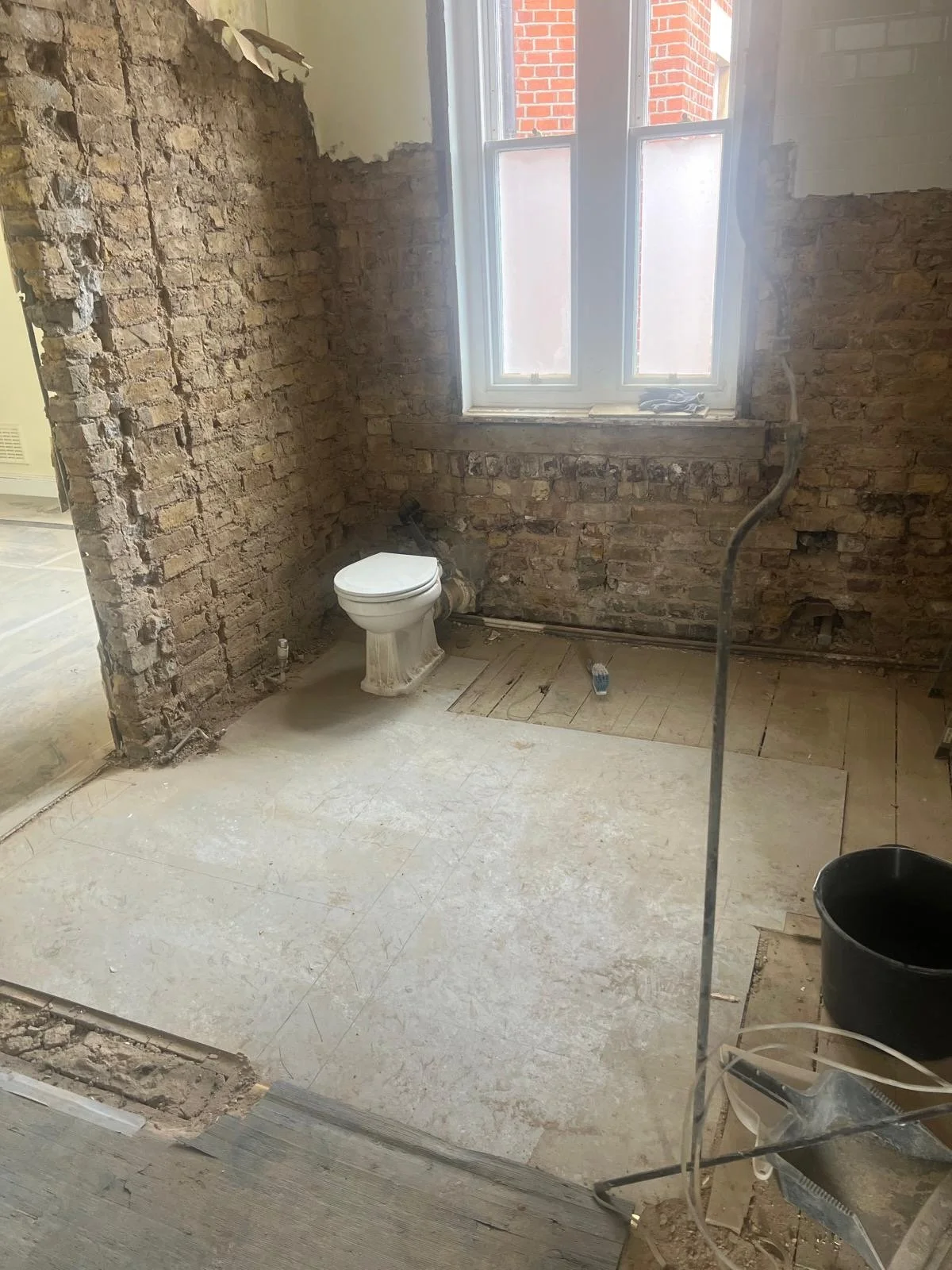 A bathroom under renovation with exposed brick walls, a toilet, a window, and construction materials scattered on the unfinished floor.