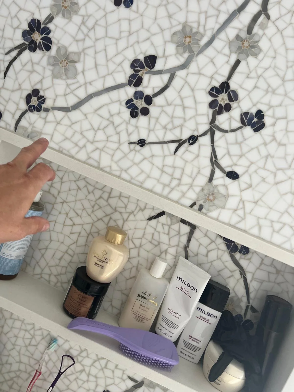 Bathroom shelf containing various hair and skincare products including a shampoo, restorative treatment, repair masks, a purple hairbrush, and other personal care items, set against a decorative floral mosaic tiled wall.