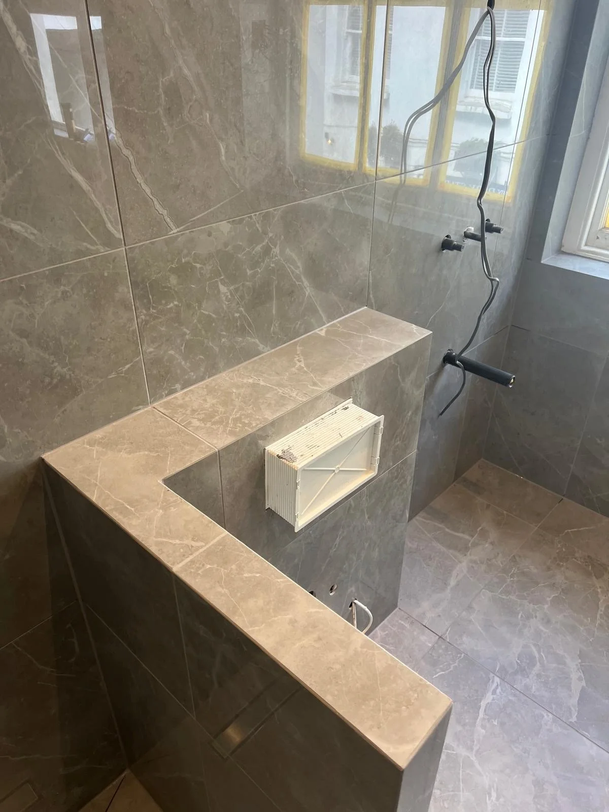 Empty shower area with grey marble tiles, a small window, busted heater, and exposed electrical wiring.