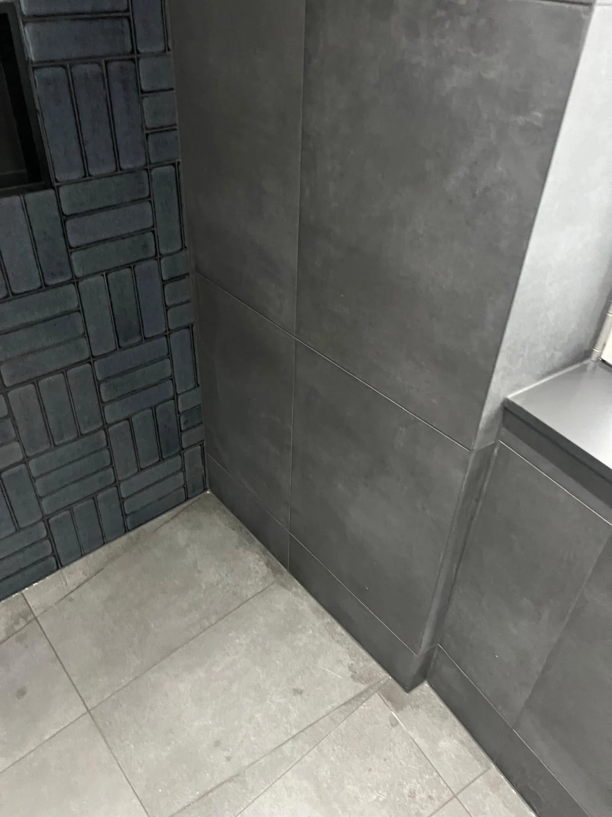 Close-up of a corner in a modern bathroom with dark gray tiles on one wall, larger gray tiles on another, and beige ceramic floor tiles.