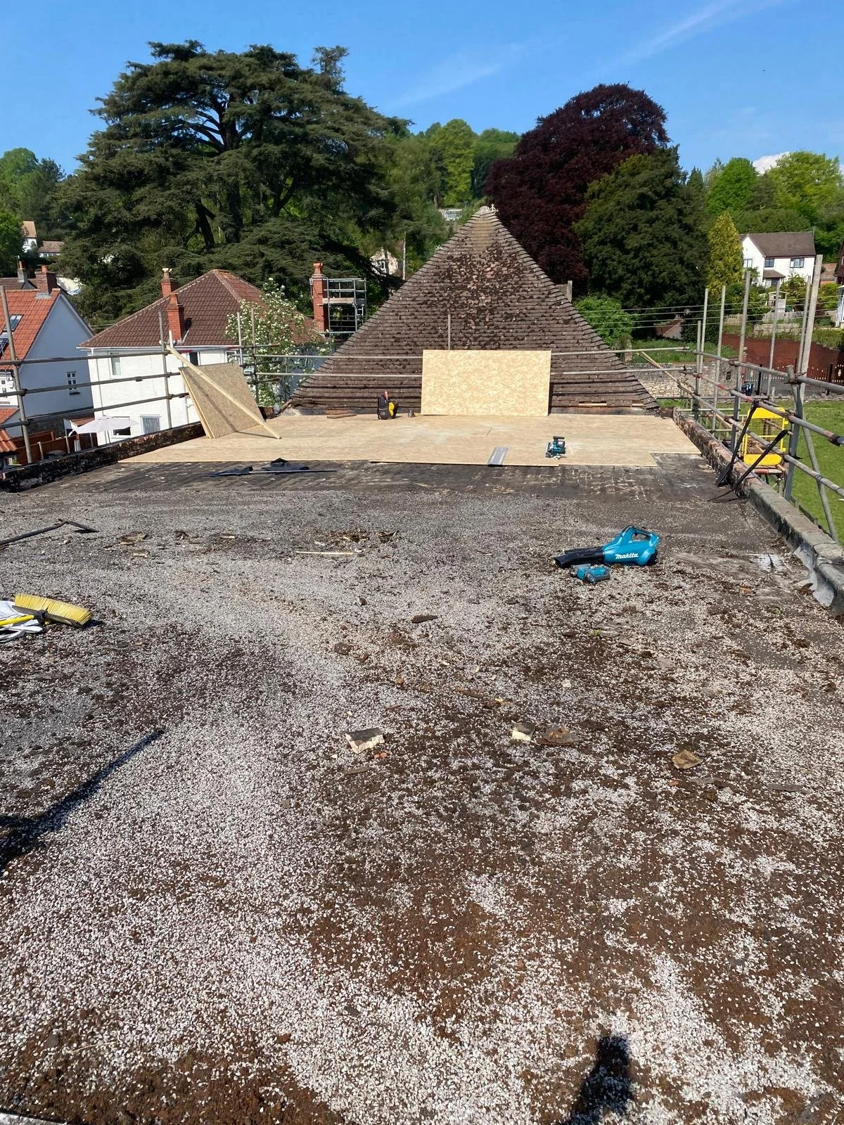 Construction site on a rooftop with construction materials, tools, and partially installed wooden platform, with trees and houses in the background.