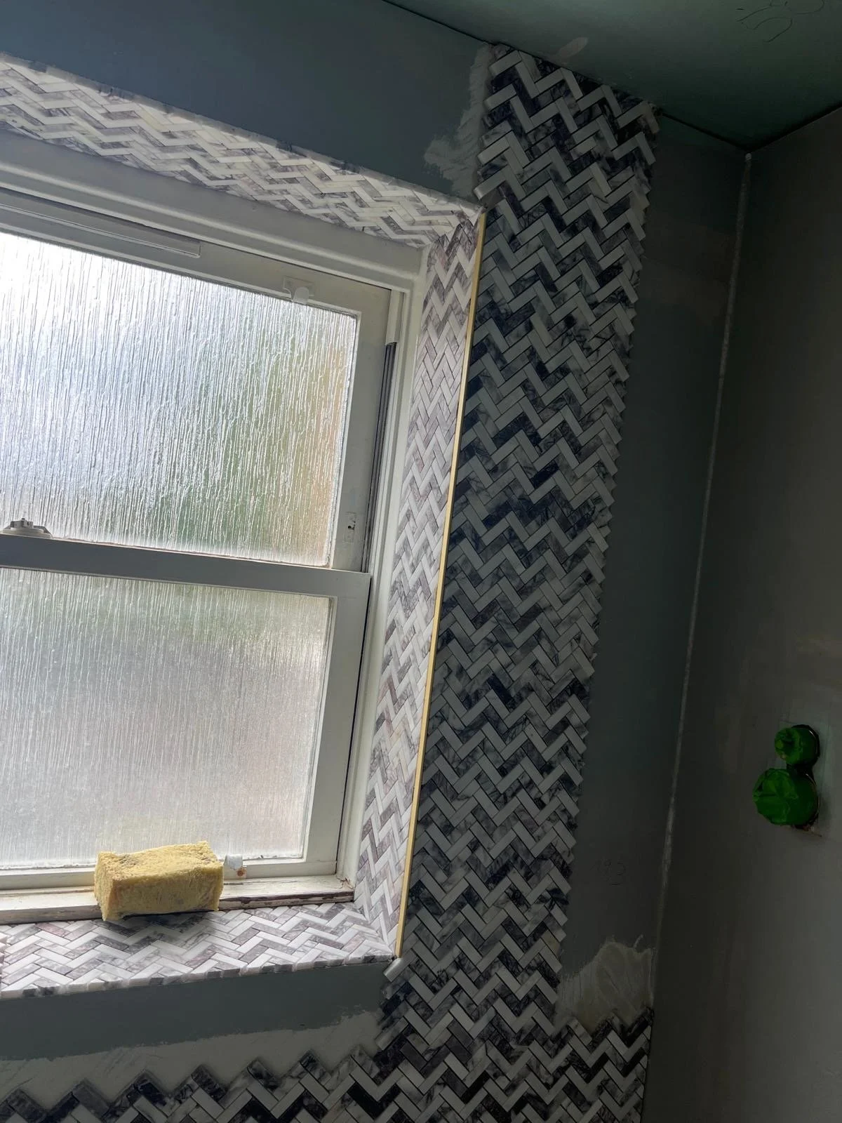 A window with textured glass panes, a yellow sponge resting on the windowsill, and decorative tile work with a herringbone pattern surrounding the window frame and extending along an adjacent wall. Green plastic bags are hanging on the wall nearby.