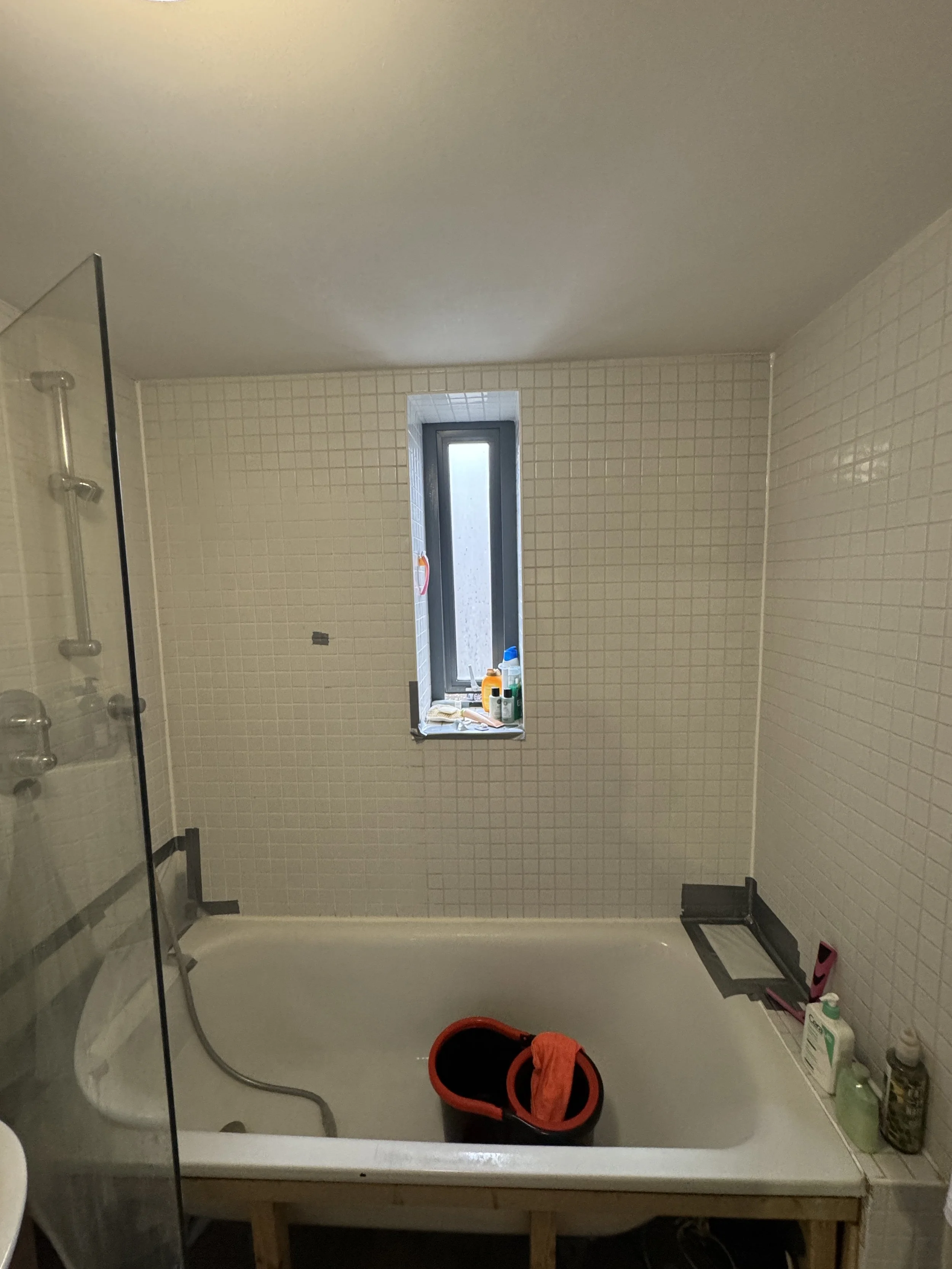 Bathroom with a small window, beige tiled walls, and a shower enclosure with a glass door. There are cleaning supplies and personal care items on the windowsill, and a small trash bin on the floor. The image is slightly tilted, showing the bathroom f
