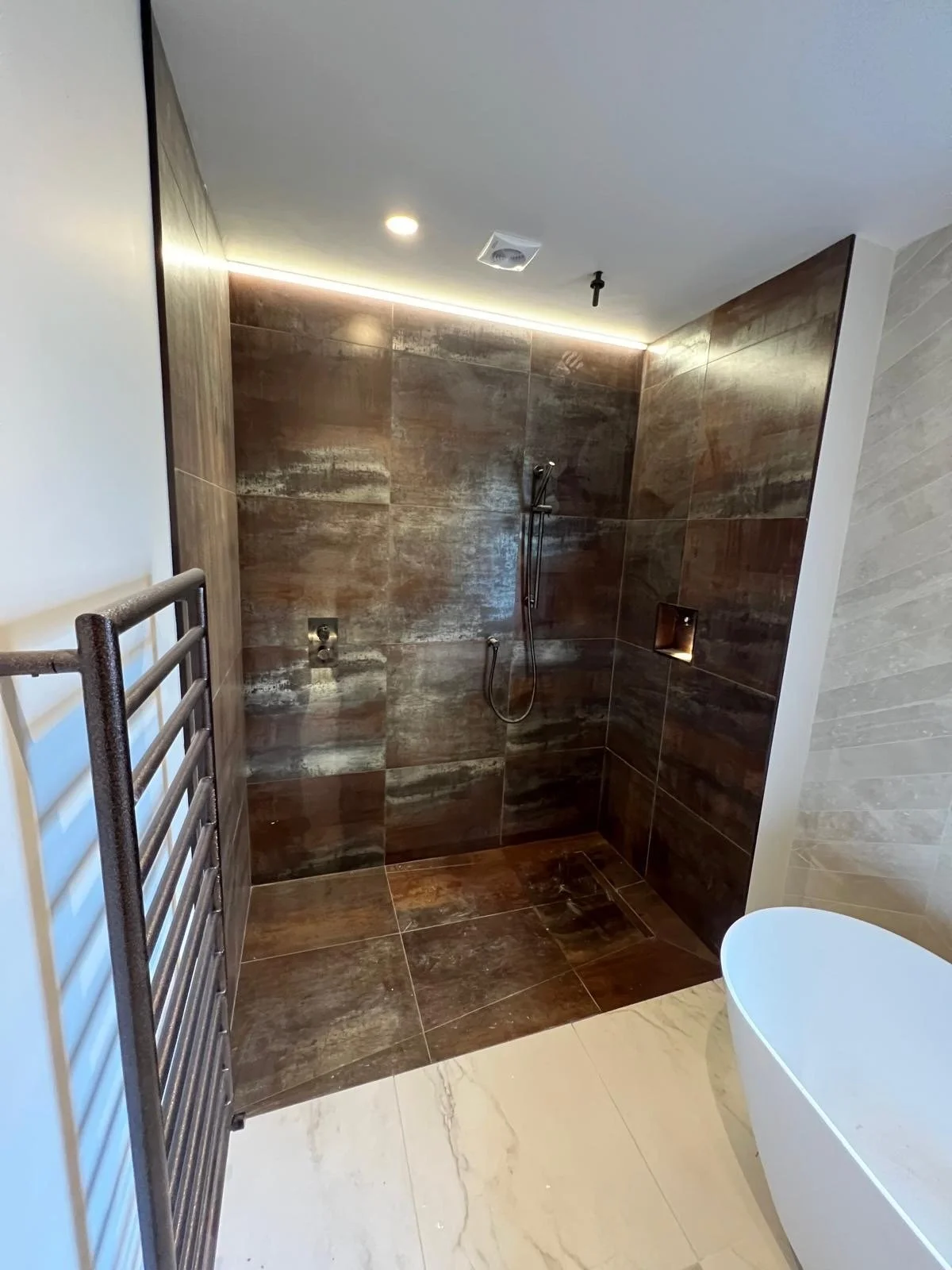 A modern bathroom with a walk-in shower featuring dark, textured tiles, a handheld showerhead, a small niche for toiletries, a heated towel rail, a white toilet, and cream-colored marble tile flooring.