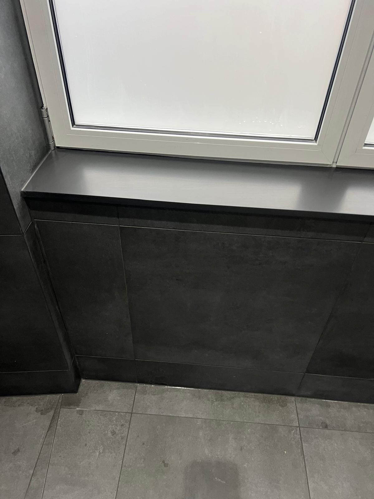View of a black tiled wall with a windowsill and frosted window, floor is gray tiles.