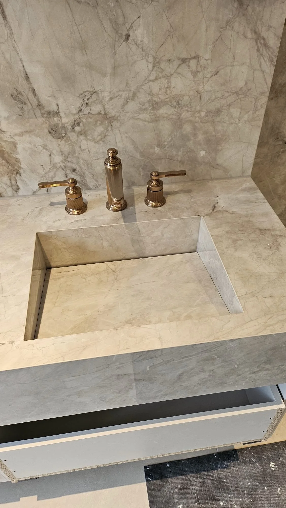 A marble bathroom sink with a rectangular basin, two gold-finished faucets, and a marble backsplash.