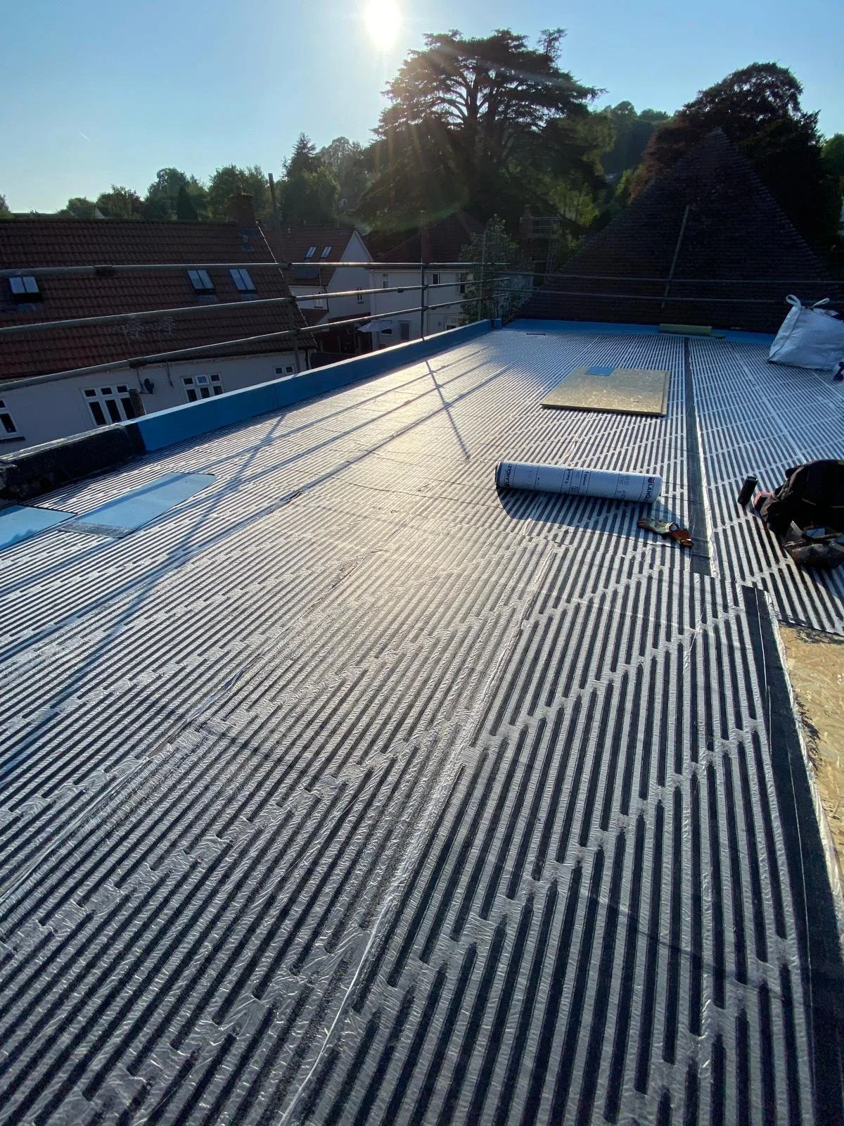 Construction site on a roof with roofing material and tools, with neighboring houses and trees in the background, and the sun shining.