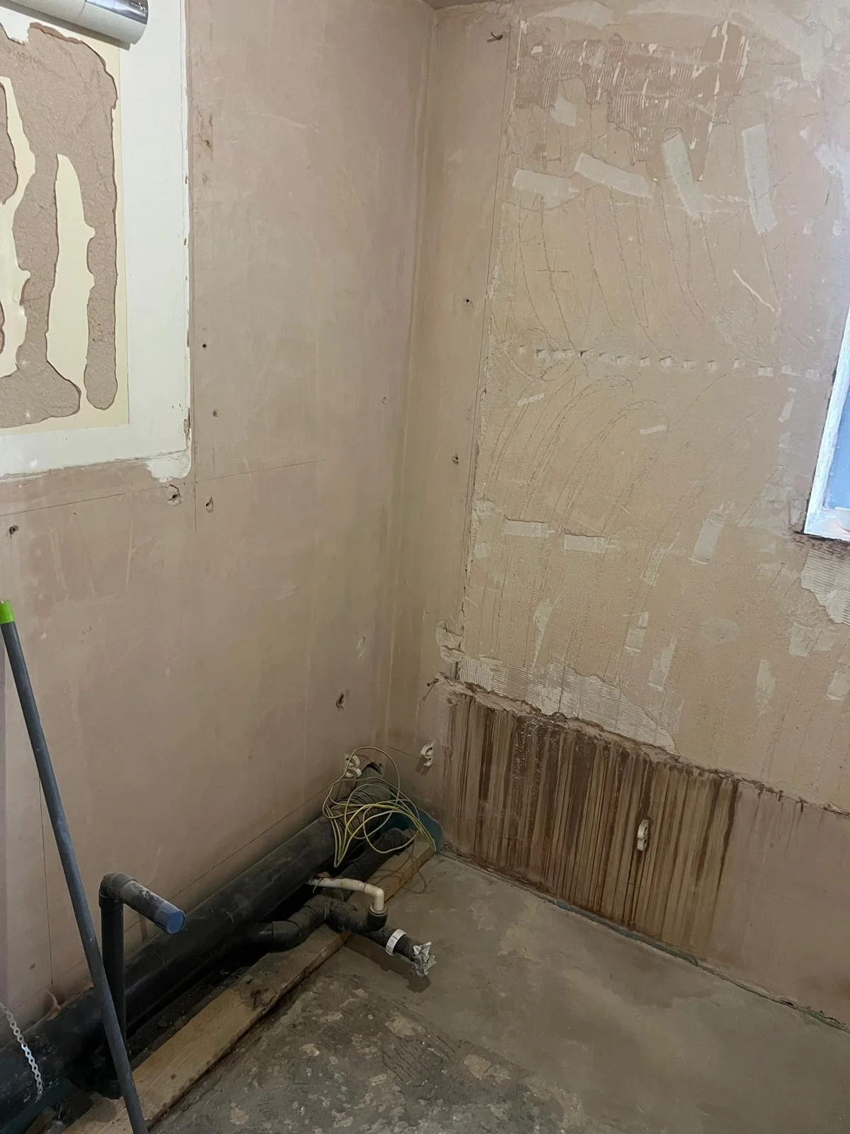 A room under construction with partially torn drywall, exposed wiring, and plumbing pipes on the floor.