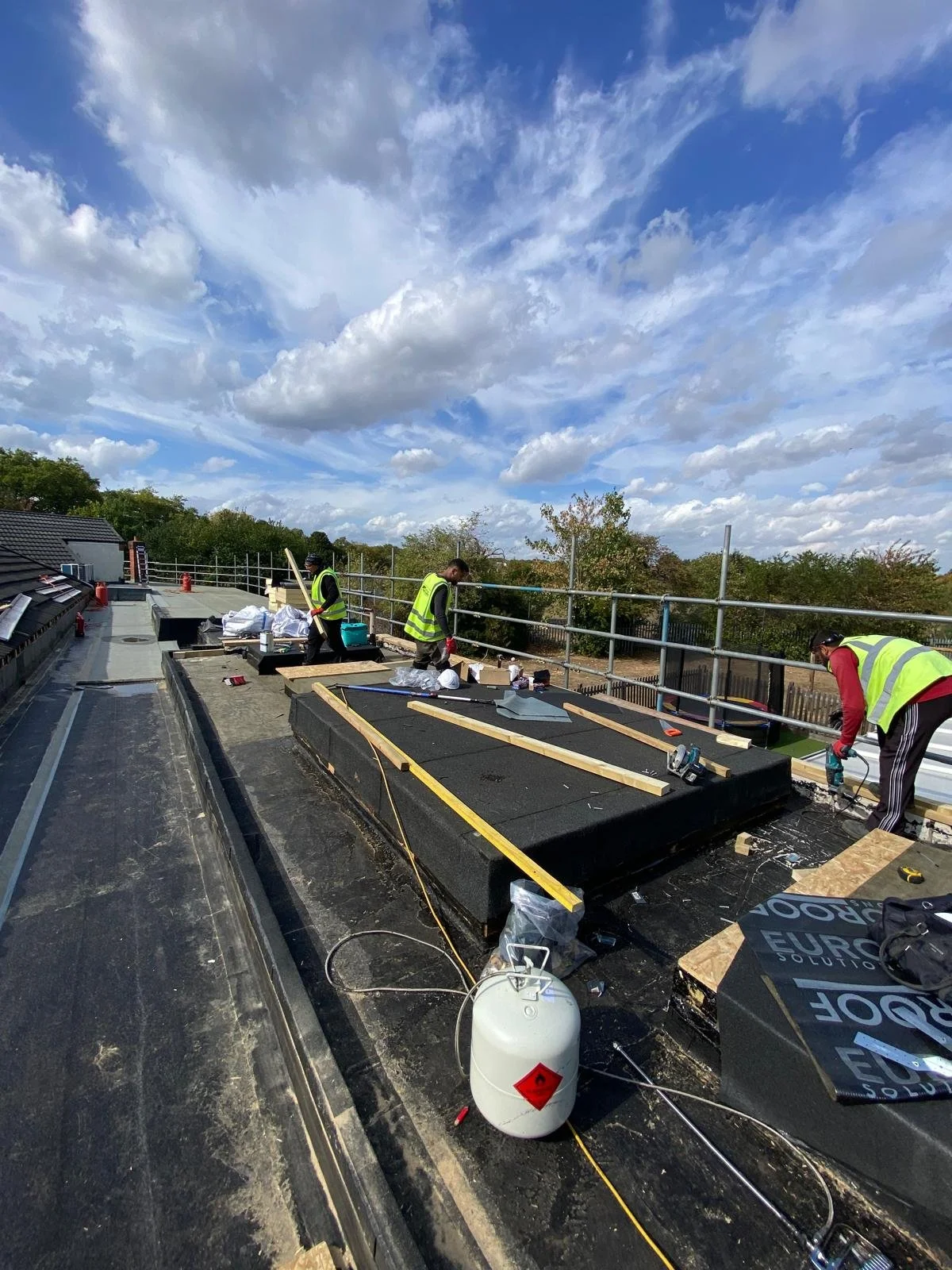 Workers installing a flat roof on a building, with tools and materials scattered around under a partly cloudy sky.
