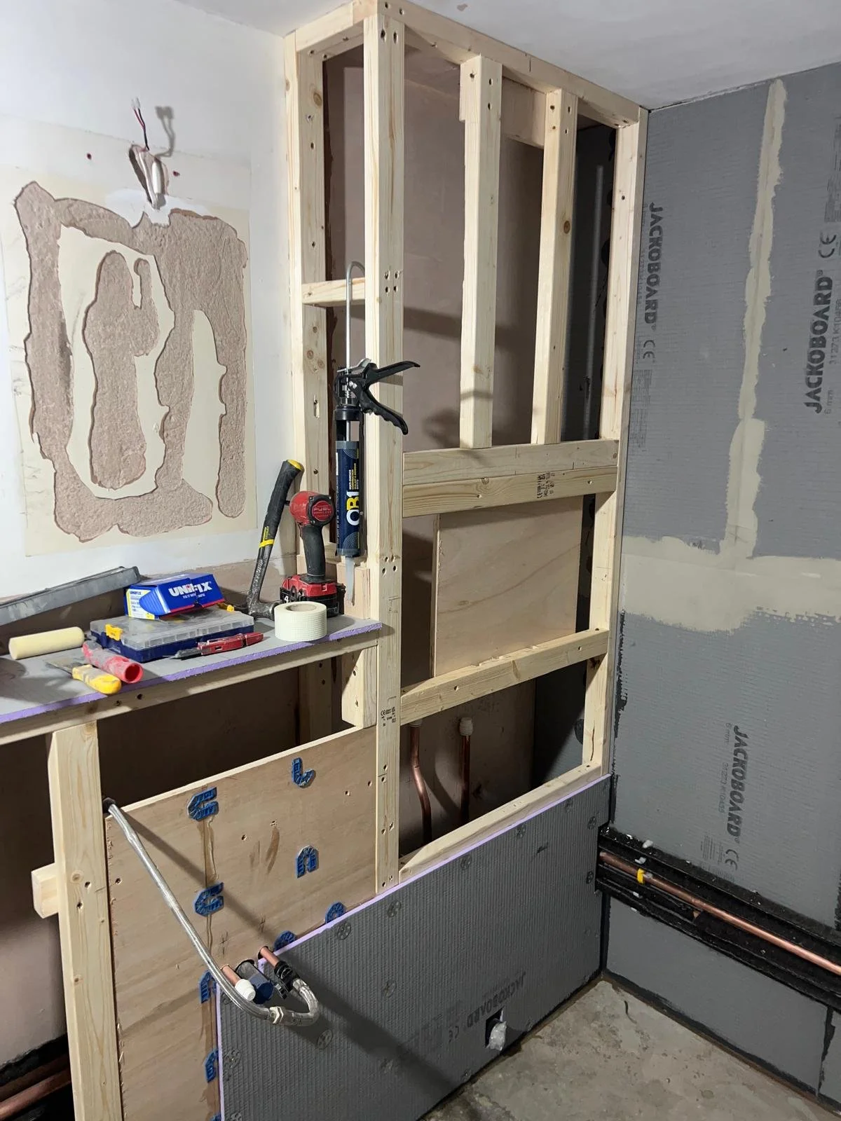 Bathroom construction site with exposed wooden framing, tools, and plumbing pipes. Part of the wall is covered with drywall and insulation.