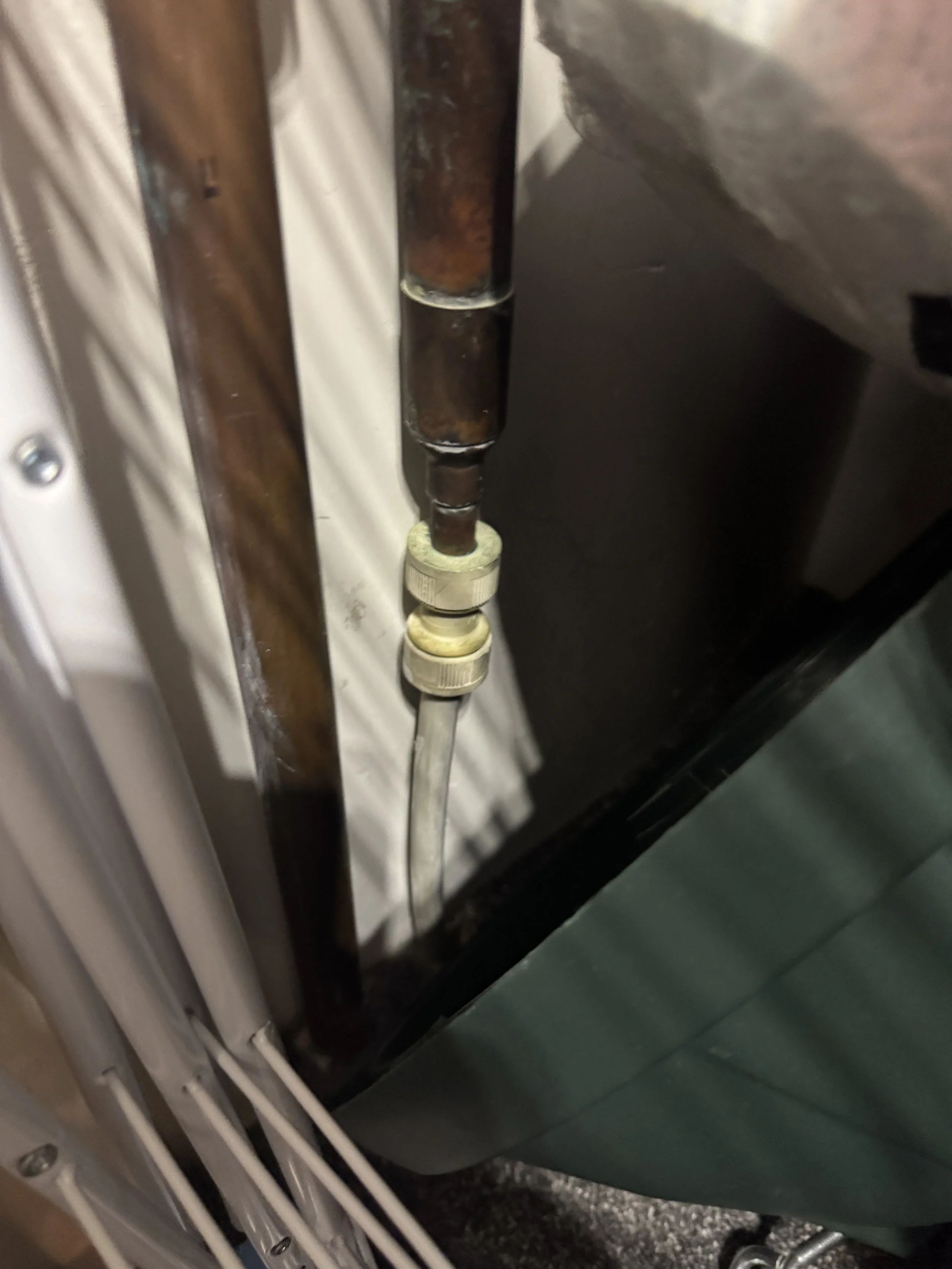 A close-up image of a rusty metallic pipe connected to a clear plastic tube with a white plastic valve at the end, located near a wooden surface and some electronic or metal components.