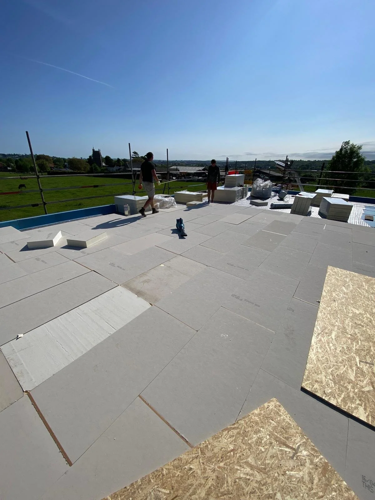 Construction workers installing large white tiles on a flat rooftop, with a rural landscape in the background under a blue sky.