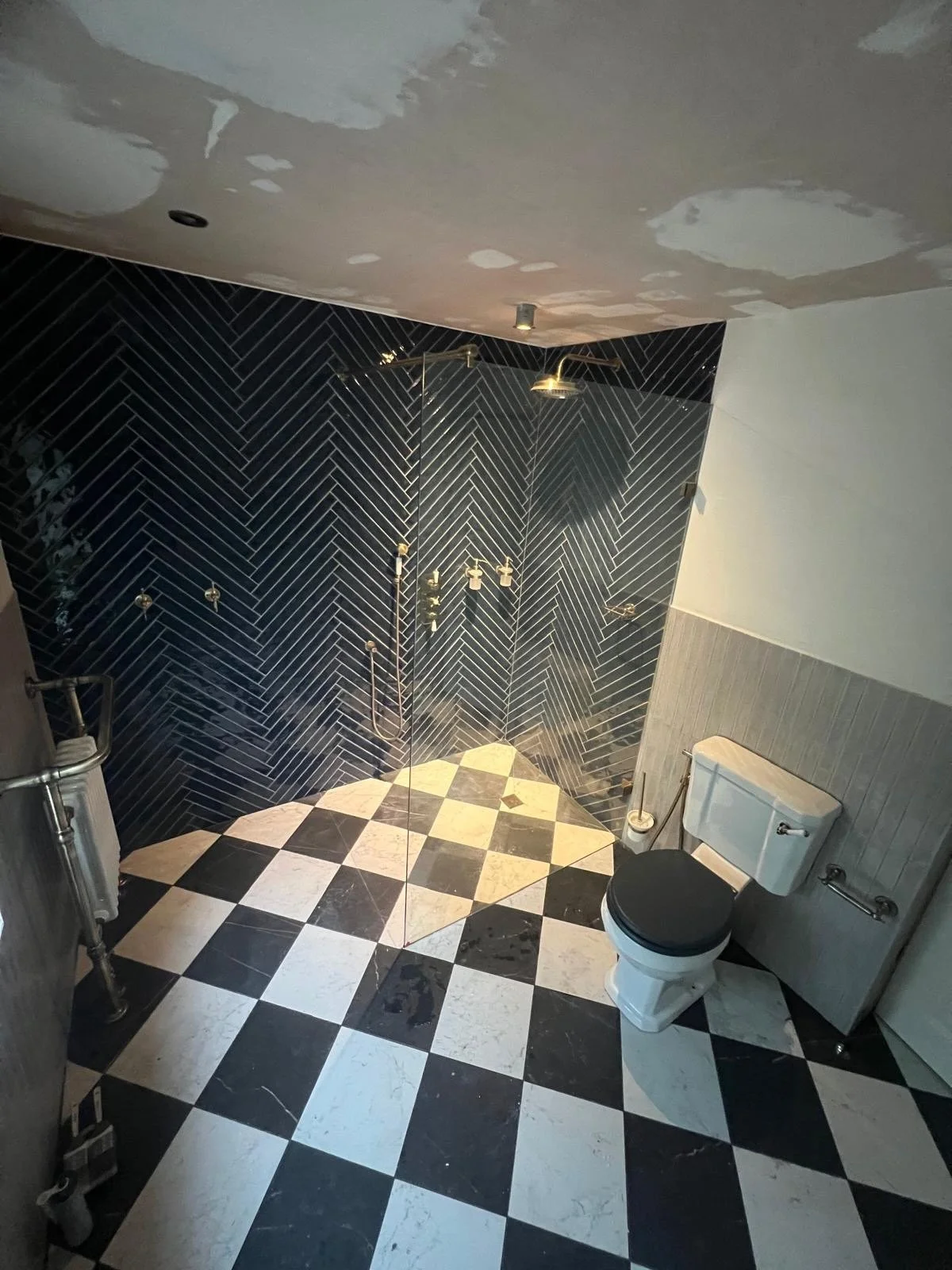 Bathroom with black and white checkered floor, black tile walls with a herringbone pattern, a white toilet with a black seat, and a shower area with brass fixtures and a glass door.