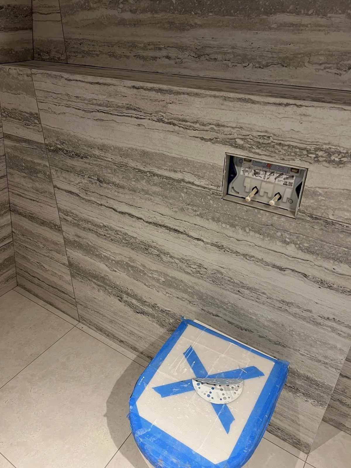 Bathroom wall with an electrical outlet box and a blue-taped stool with a metal plate on top, possibly for a sink installation.