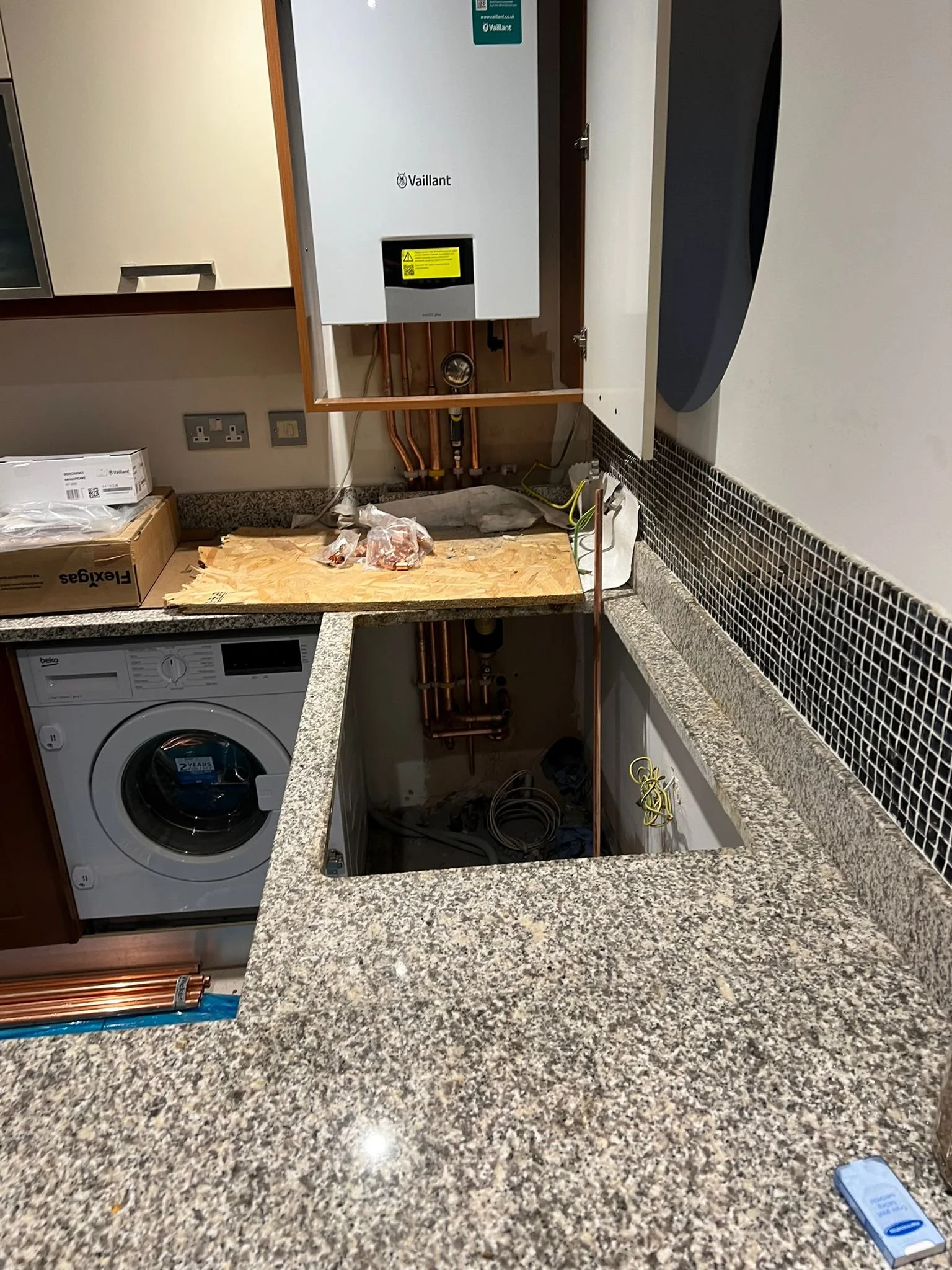 Kitchen countertop with open cabinet, electrical outlet, washing machine, and plumbing for a sink under construction. There are copper pipes, construction materials, and tools present.