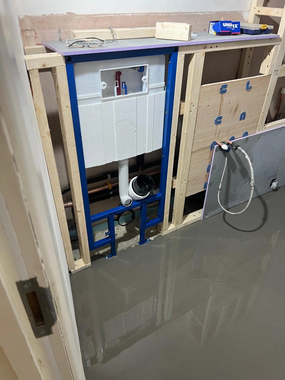 Under construction bathroom with a toilet tank frame mounted on wooden studs, plumbing pipes, and a mounted heater or radiator on the wall, with tools and materials on a countertop.
