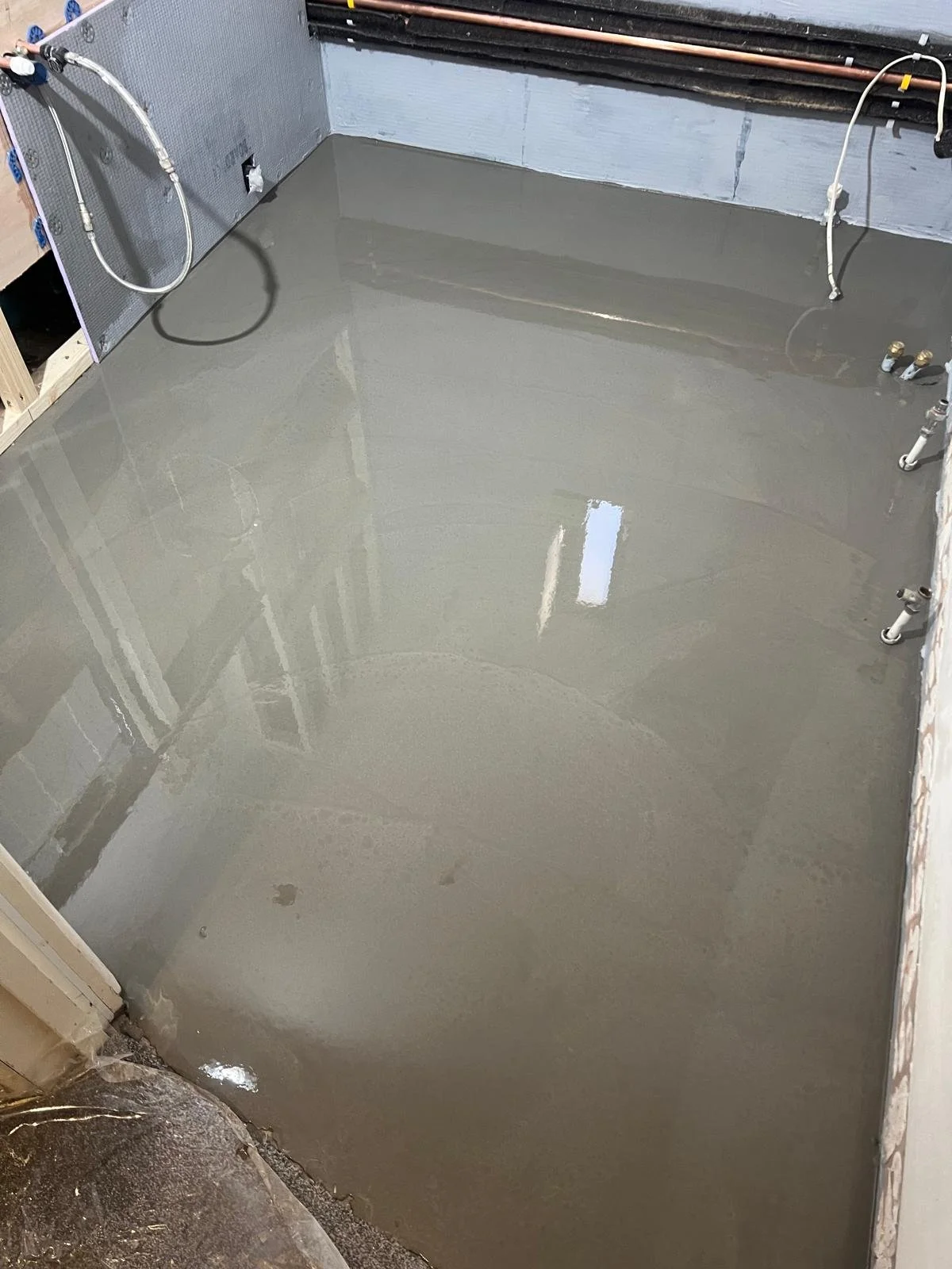 Freshly poured concrete floor in a construction site, with visible plumbing and electrical wiring.
