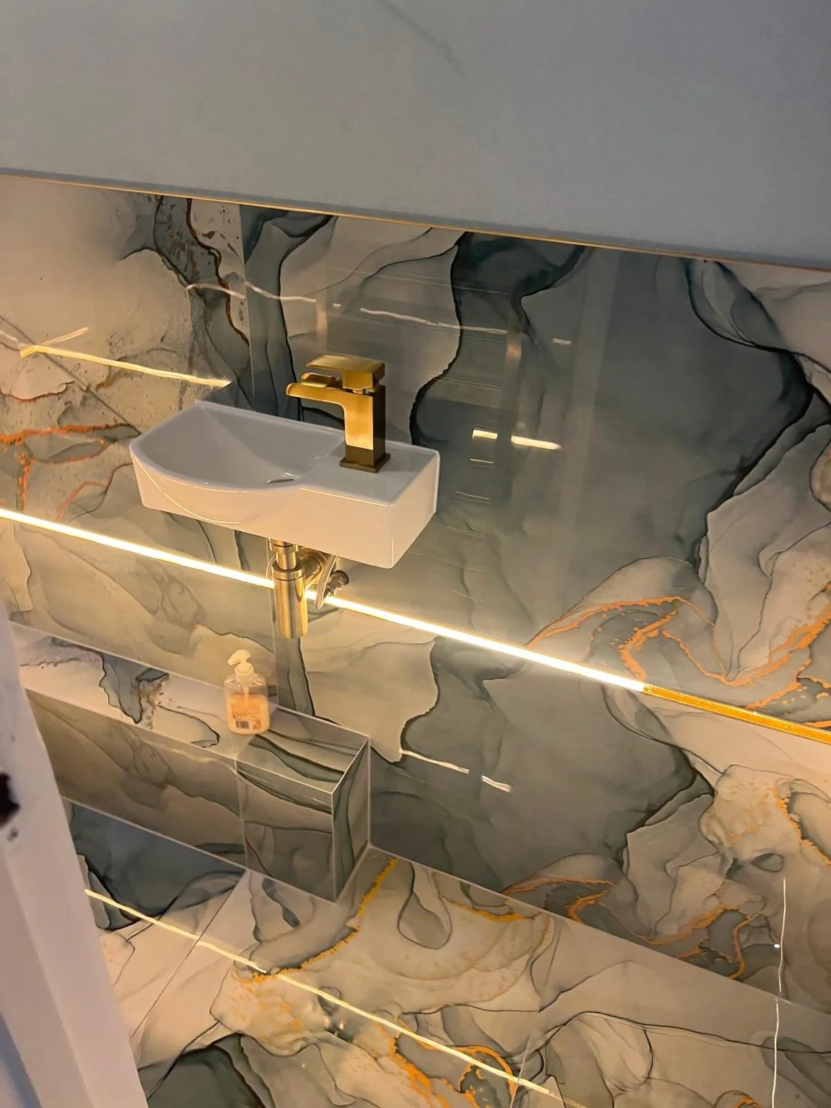 Modern bathroom with marble wall tiles, a small white sink with a gold faucet, a bottle of hand soap, and a rectangular mirror with LED lighting.