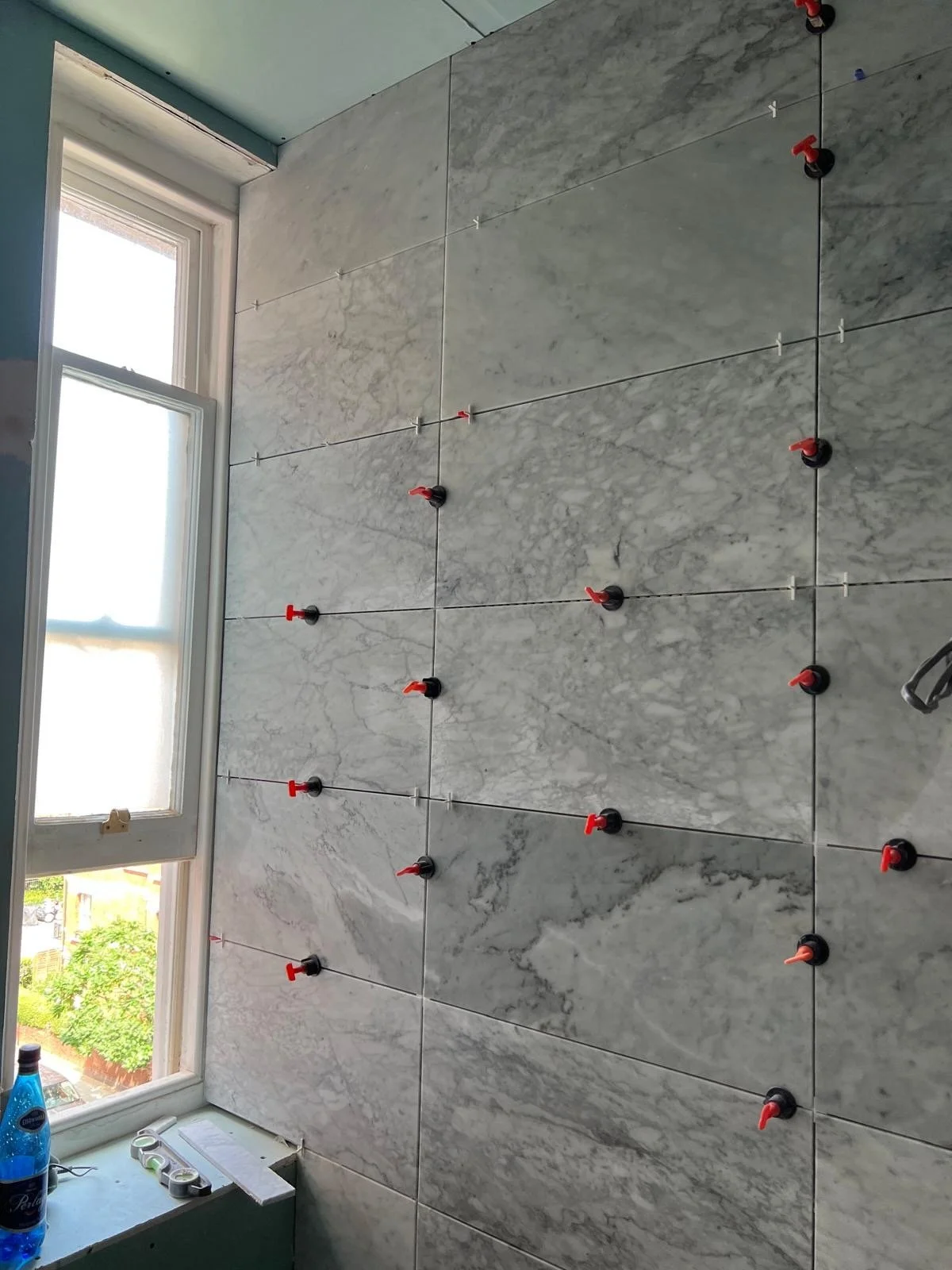 Installing tiles on a wall with spacers and clips near a window in a room, possibly during renovation.