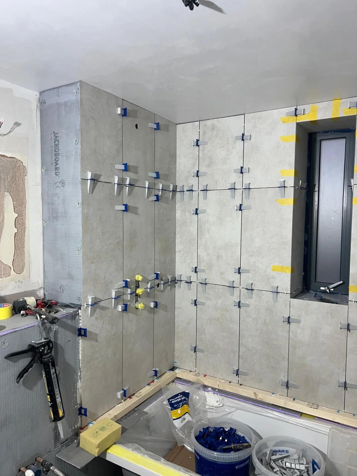 Interior wall under construction with tiles being installed, with tile spacers and tools visible.