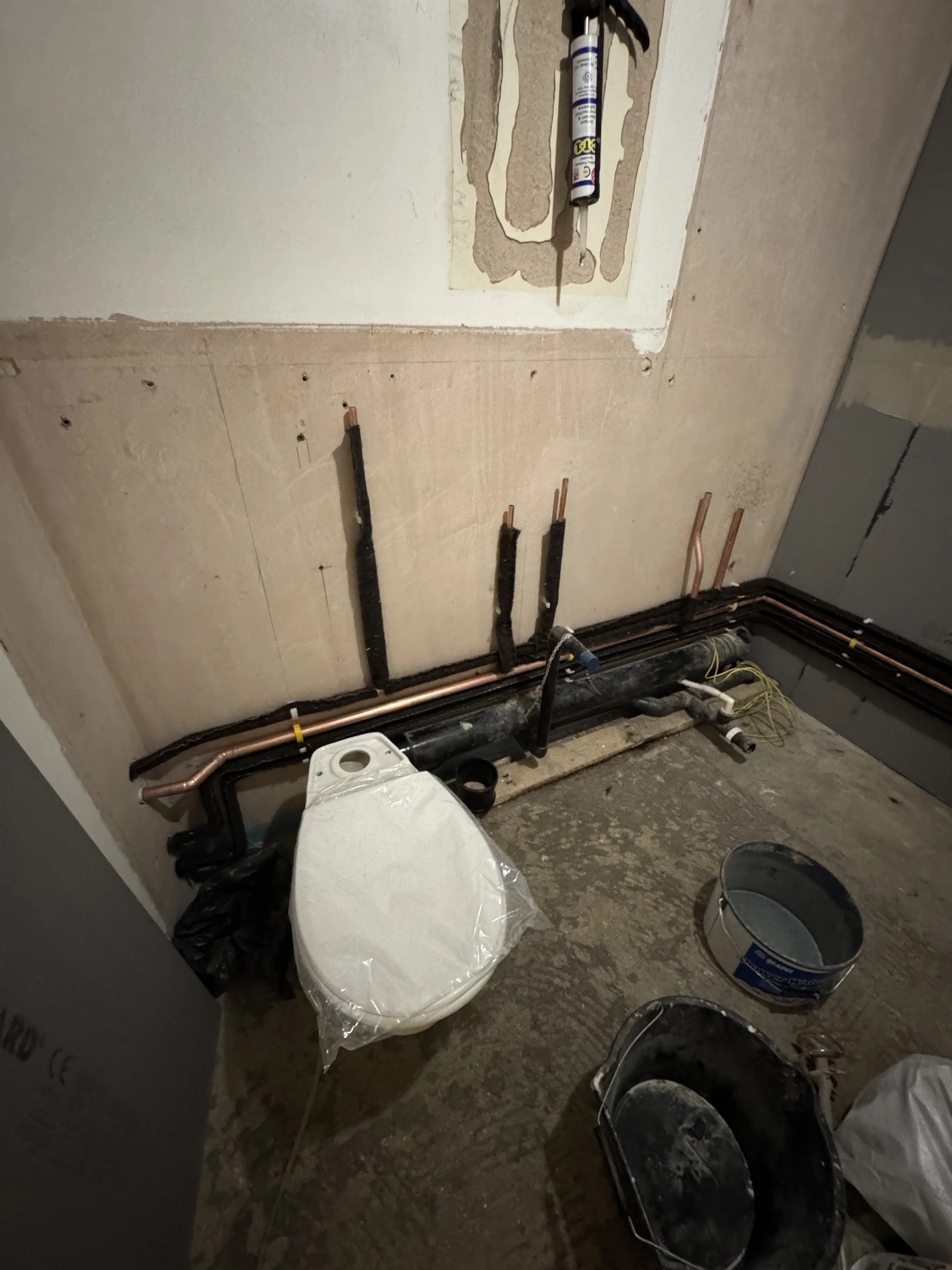 Bathroom renovation with pipes and construction materials, including a toilet wrapped in plastic, buckets, and plumbing pipes on the floor.