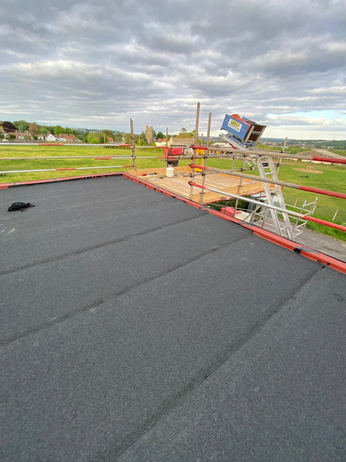 Roof with black shingles, construction scaffolding and a ladder, a wooden platform, and a cloudy sky with a rural landscape in the background.