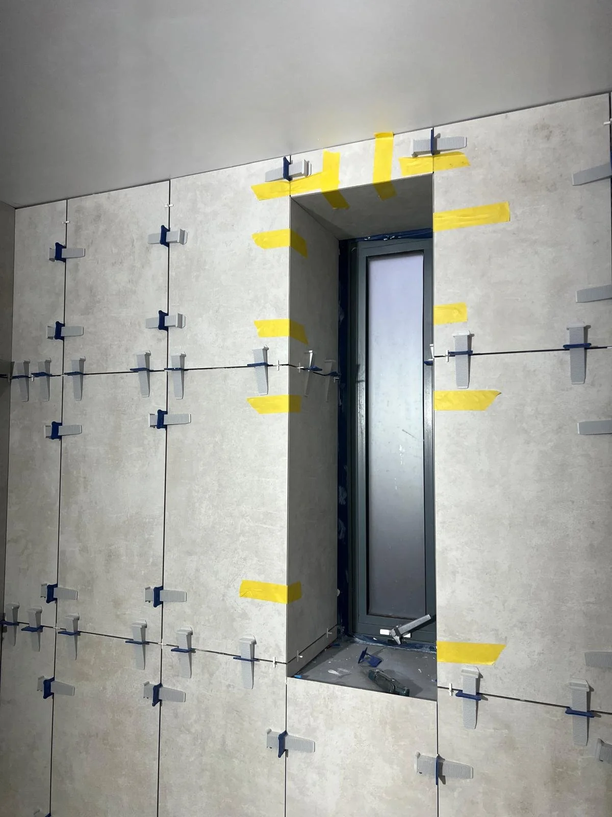 Wall with gray tiles installed and tile spacers and yellow painter's tape around a window opening during construction.