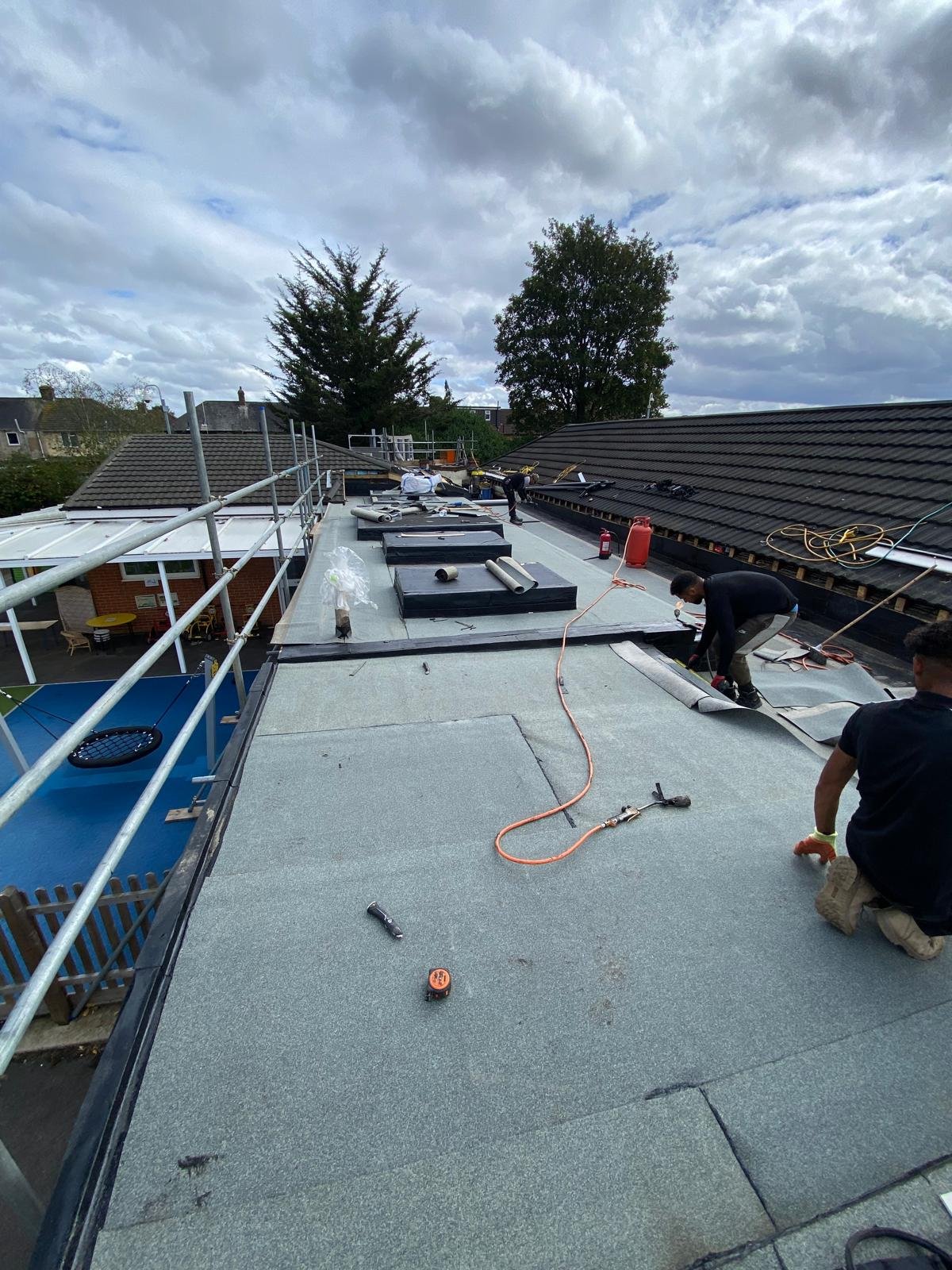 Workers installing or repairing a flat roof on a building, with tools and materials scattered around on the roof. The sky is cloudy and trees are visible in the background.