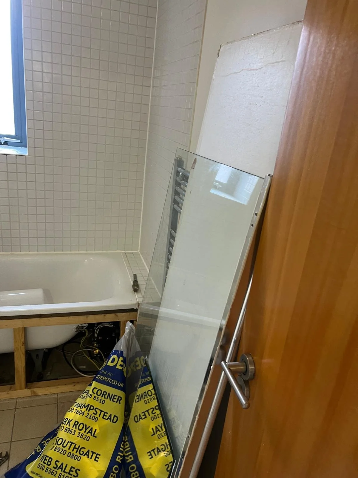 A bathroom under renovation with a partially installed bathtub, a mirror leaning against the wall, a window, and a bag from a hardware store on the floor.