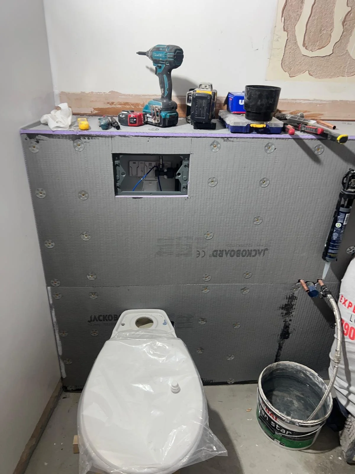 Bathroom under renovation showing a toilet with a plastic cover, construction tools, and materials like drywall, a drill, a bucket, and a wall with partially finished drywall.