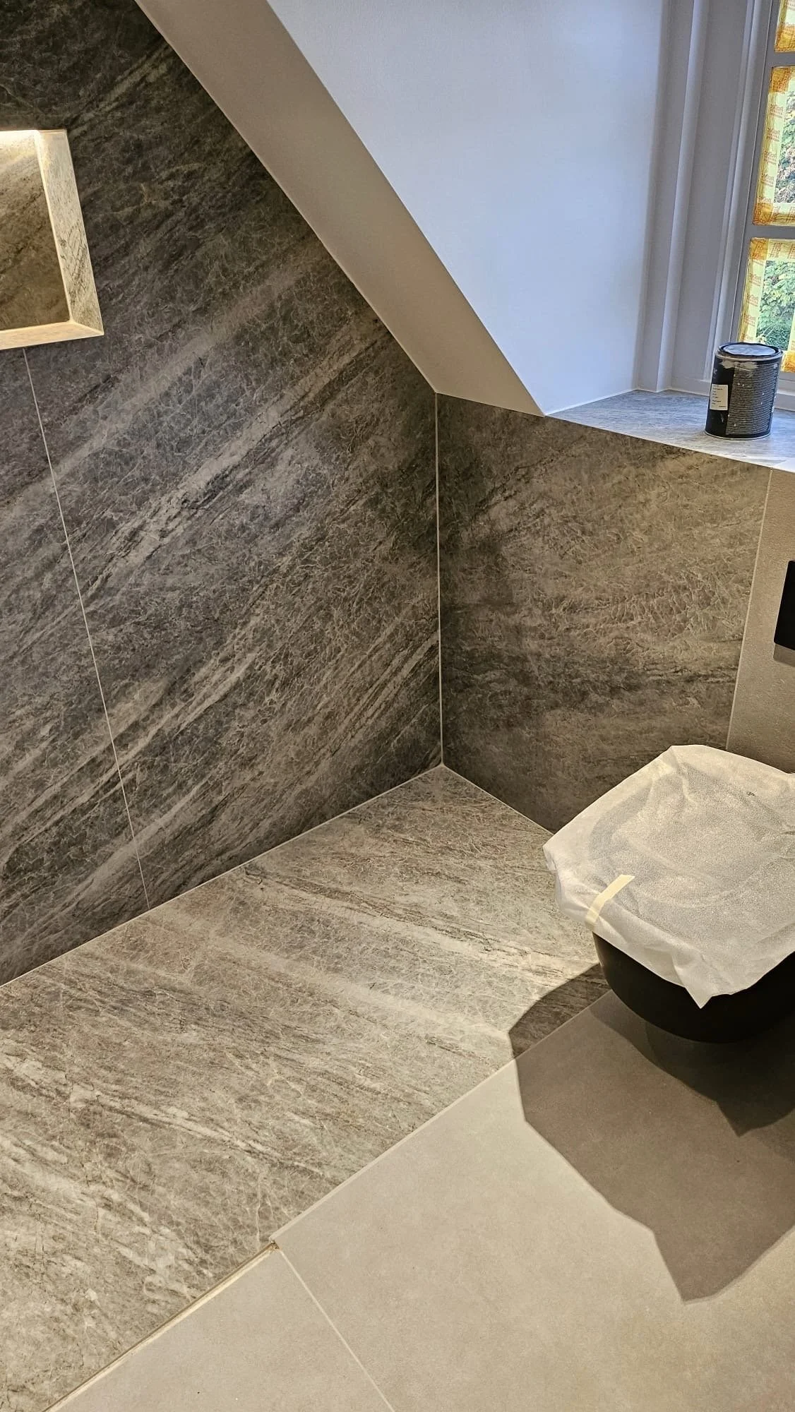 A bathroom corner with brown textured tiles on the wall and a white tiled floor. There is a small window with yellow patterned curtains, a black can on the window ledge, and a black toilet with a white cover.