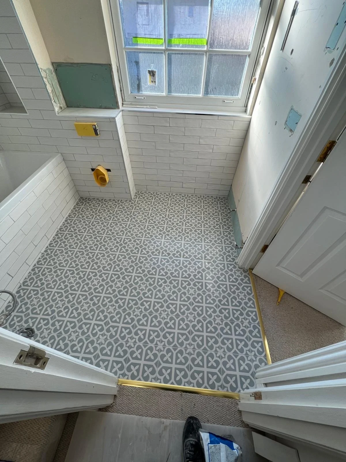 A room under renovation with patterned gray and white floor tiles, white subway tile walls, an empty window, and unfinished wall areas with construction materials and tools around.