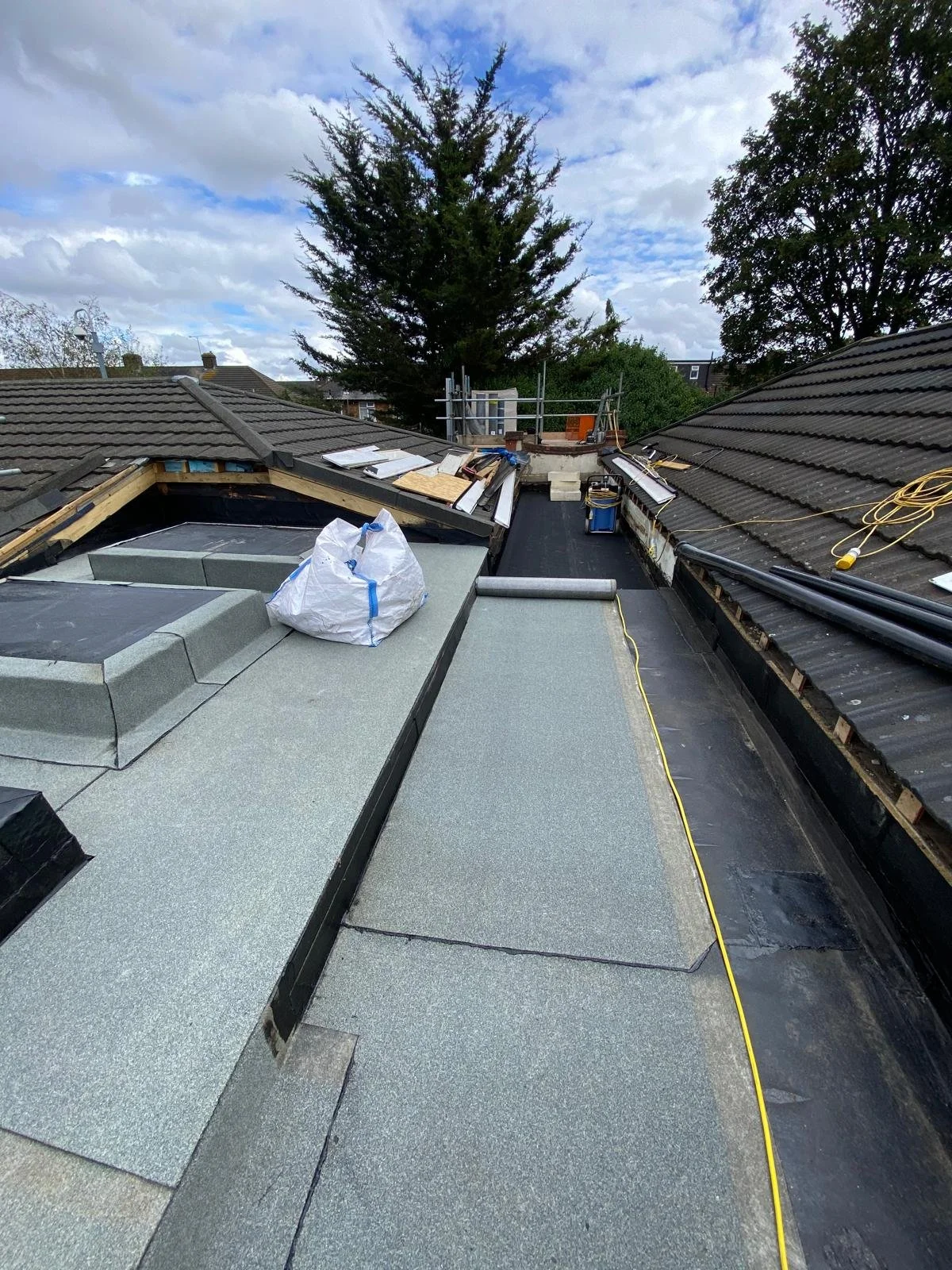 Rooftop under construction with new roofing material, tools, and materials stored on the surface. Trees and a partly cloudy sky in the background.