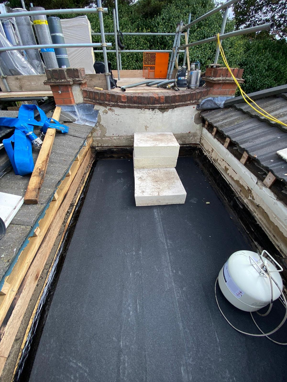 Roof construction with a black waterproofing layer, two stacked concrete blocks, construction tools, and materials visible.