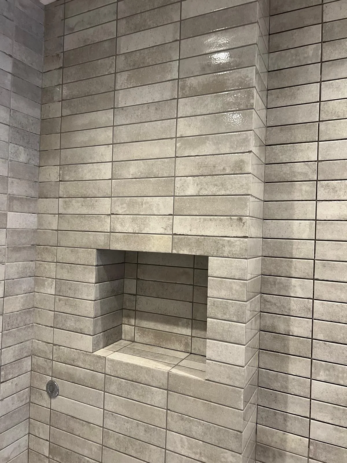 A corner of a tiled shower with beige rectangular tiles. There is a built-in shelf open to the shower area with the same tiles. A circular drain is visible in the lower left corner.
