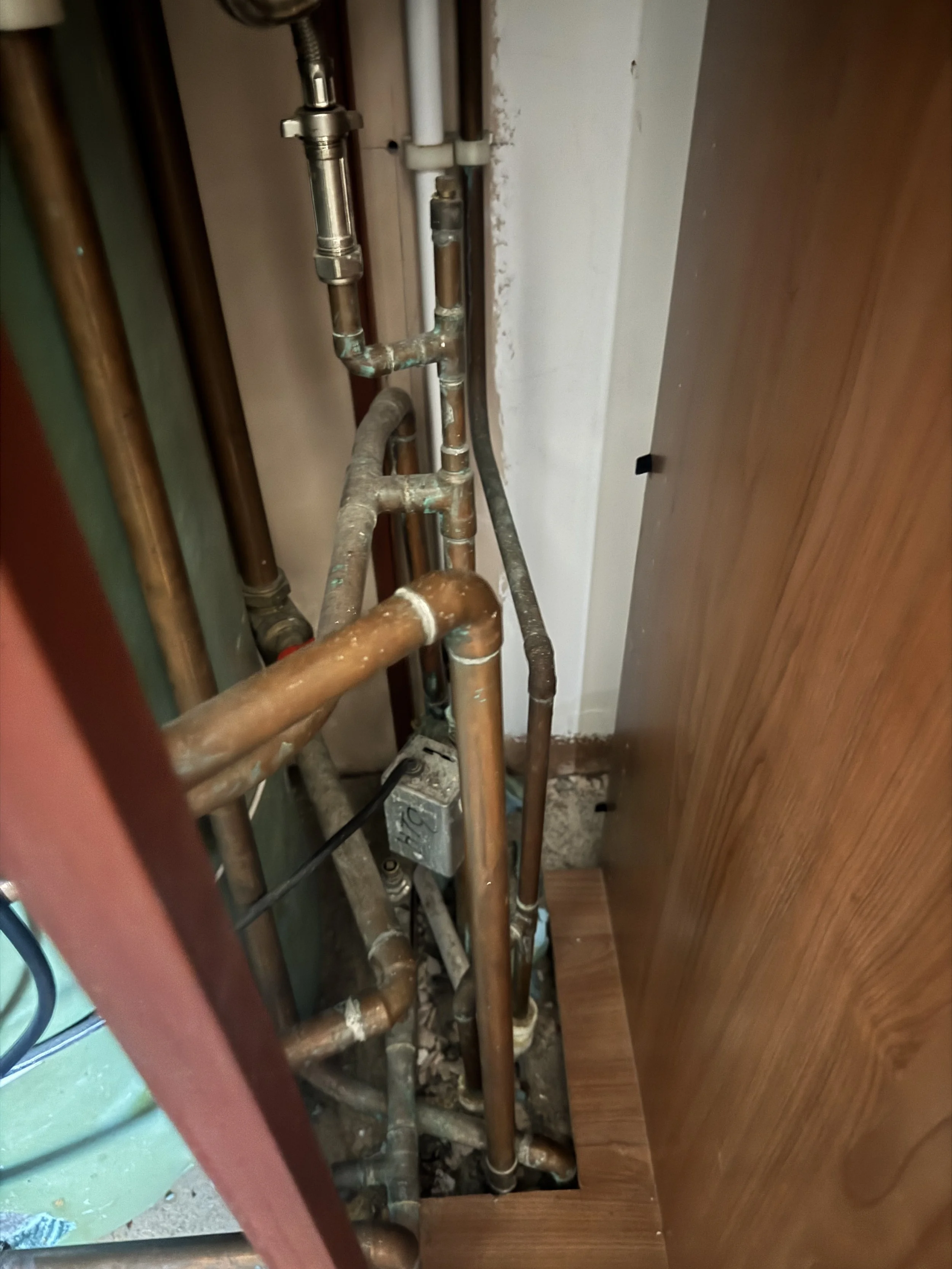 A view of various copper and metal pipes in a confined space, likely under a sink or appliance, with some white plastic pipes and an electrical component visible at the back.
