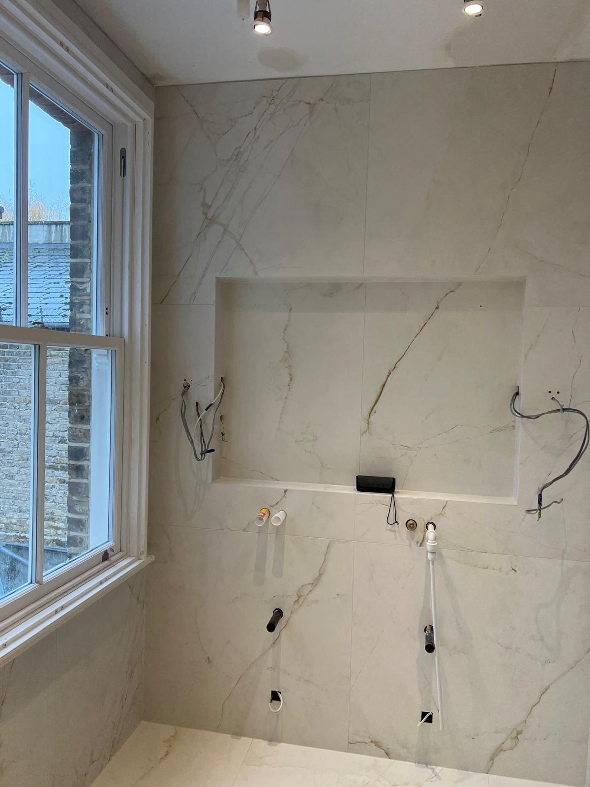 A bathroom under construction with marble wall tiles, exposed plumbing, electrical wiring, and a window showing neighboring houses.
