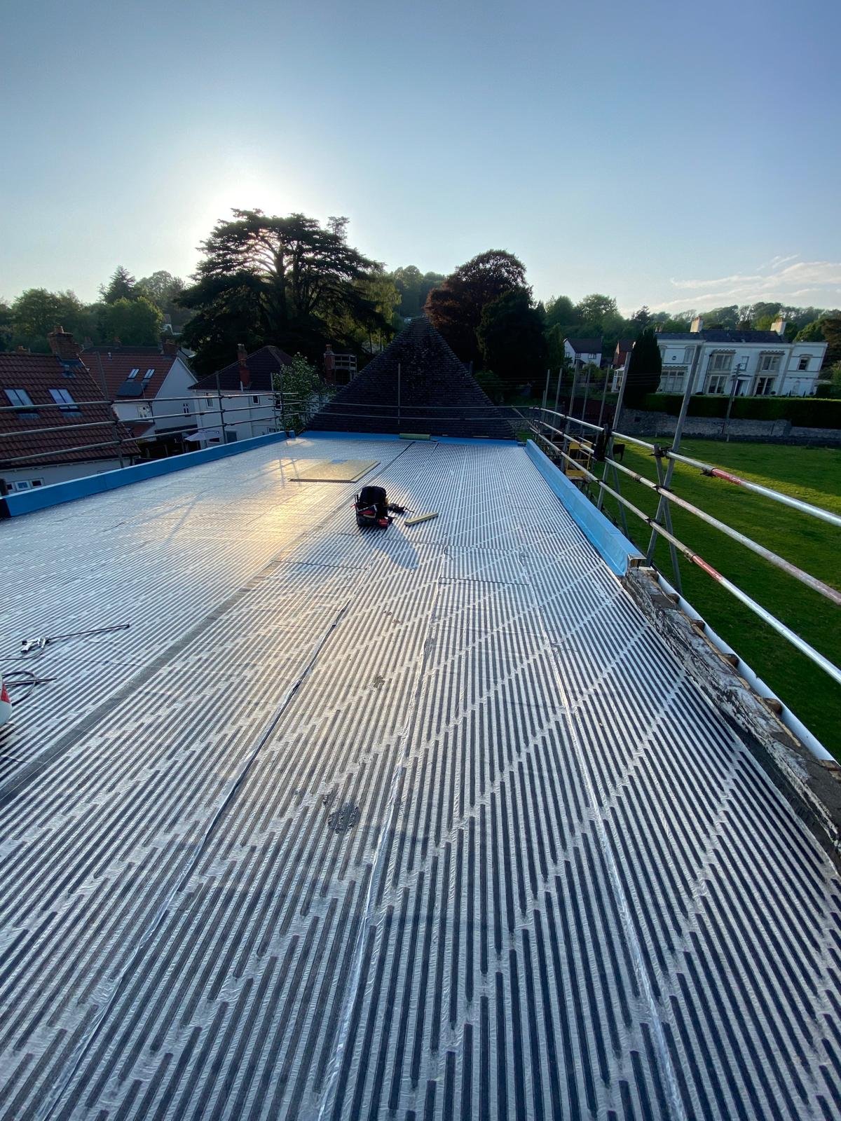 Construction scaffolding and aluminum roofing panels on a building, with tools and equipment lying on the roof, surrounded by trees and houses, during sunset.