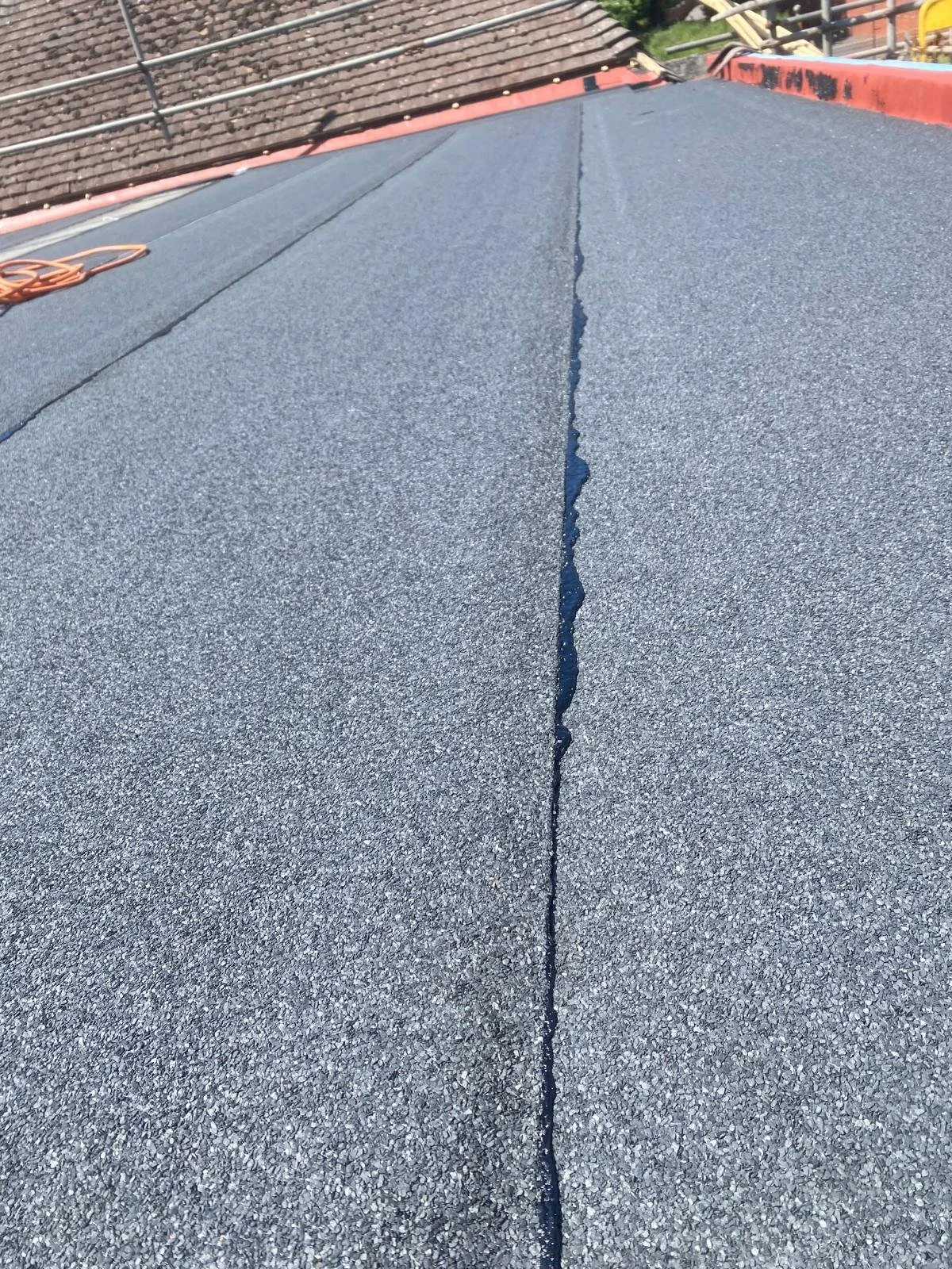 Close-up of a roof with a long crack running down the asphalt shingles, with other rooftops and trees visible in the background.