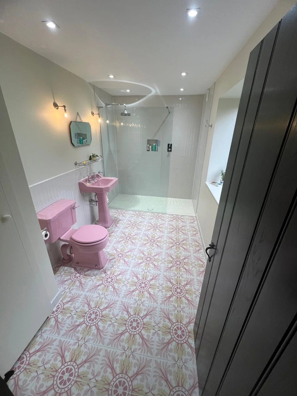 Bathroom with pink toilet and sink, patterned tile floor, walk-in shower area with glass enclosure, wall-mounted shelves, and a window ledge with a small potted plant.