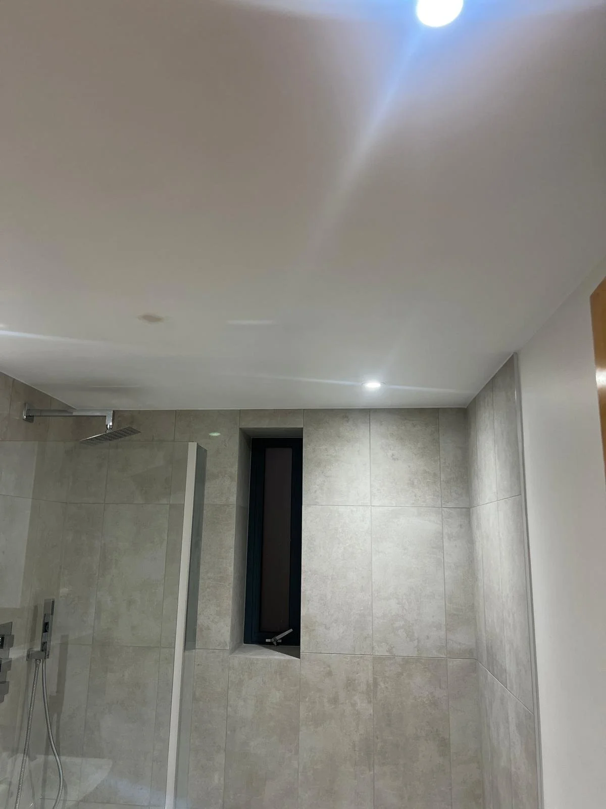 A modern bathroom with beige tiled walls, a shower area with a showerhead, a tall narrow window, and ceiling lights.