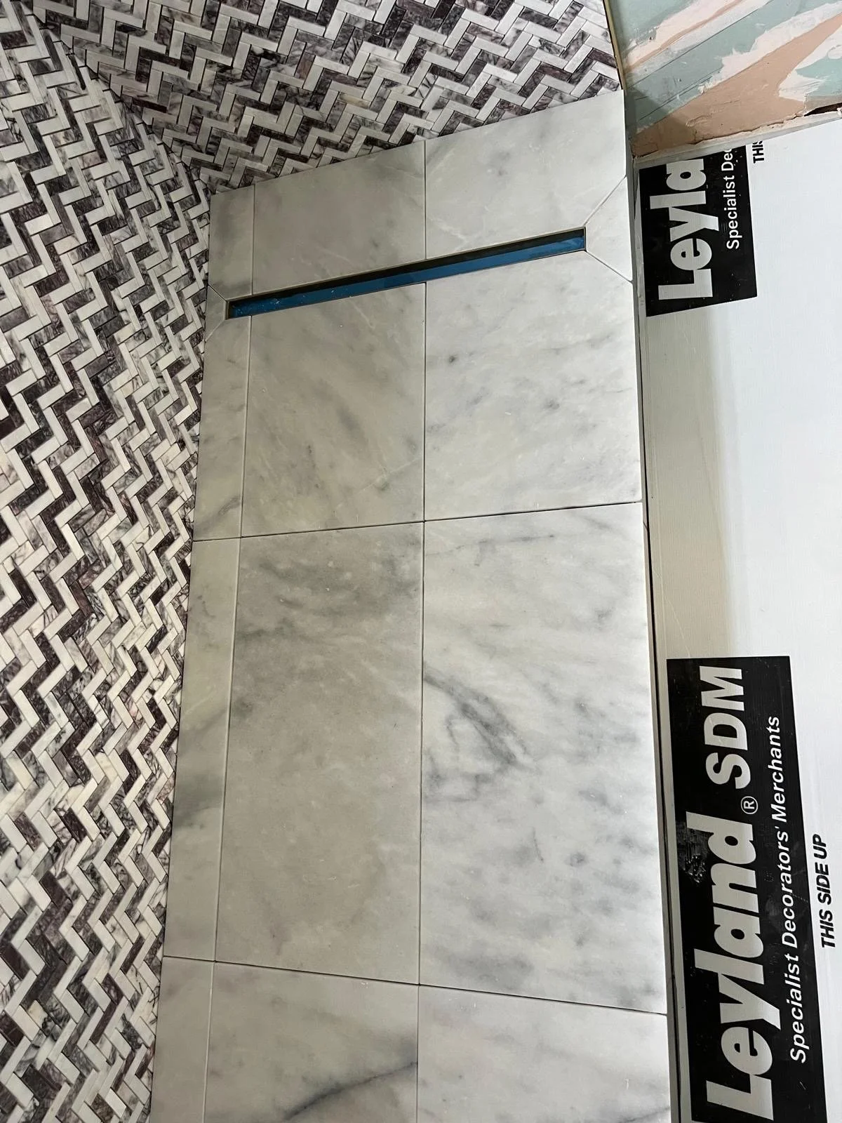 Tile flooring with large light gray marble tiles and a decorative wall with black, white, and gray geometric mosaic tiles, with a partially completed installation and construction signage.