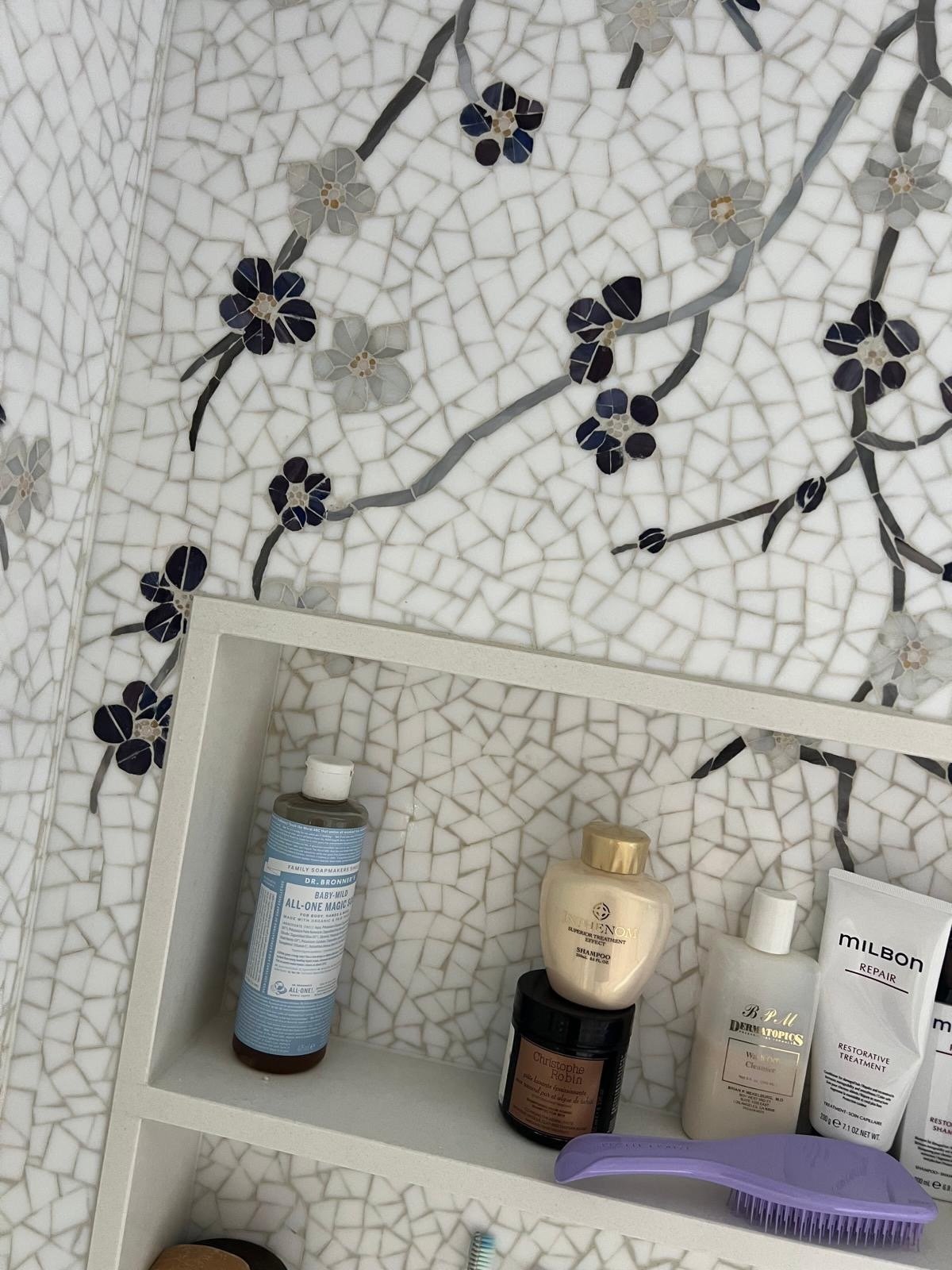 Bathroom shelf with skincare products, shampoo, conditioner, and a purple hairbrush in front of a floral mosaic wall.