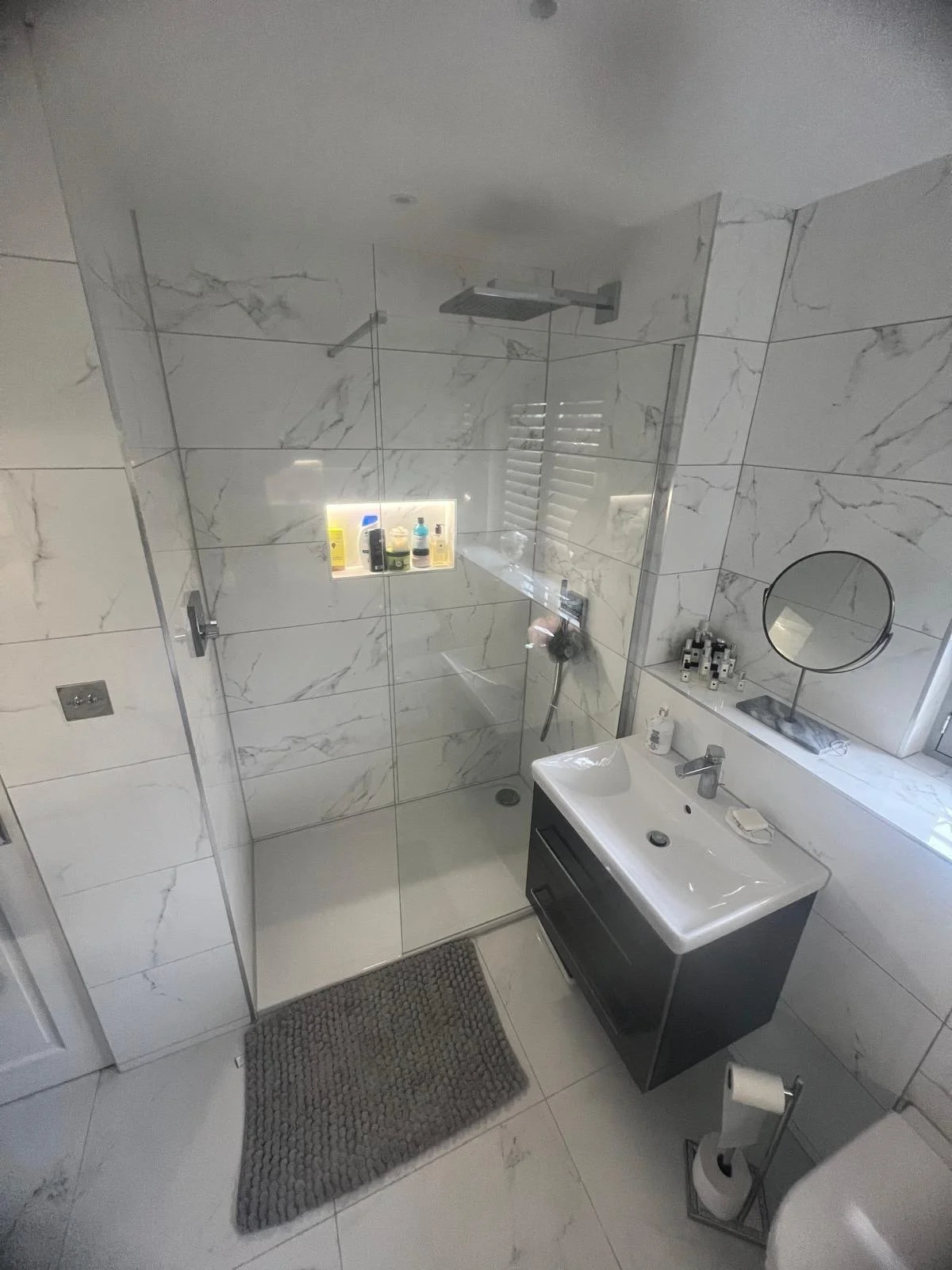 A modern bathroom with a glass-enclosed shower, marble walls, a window with blinds, a sink with a dark vanity, a mirror, and various toiletries and accessories.