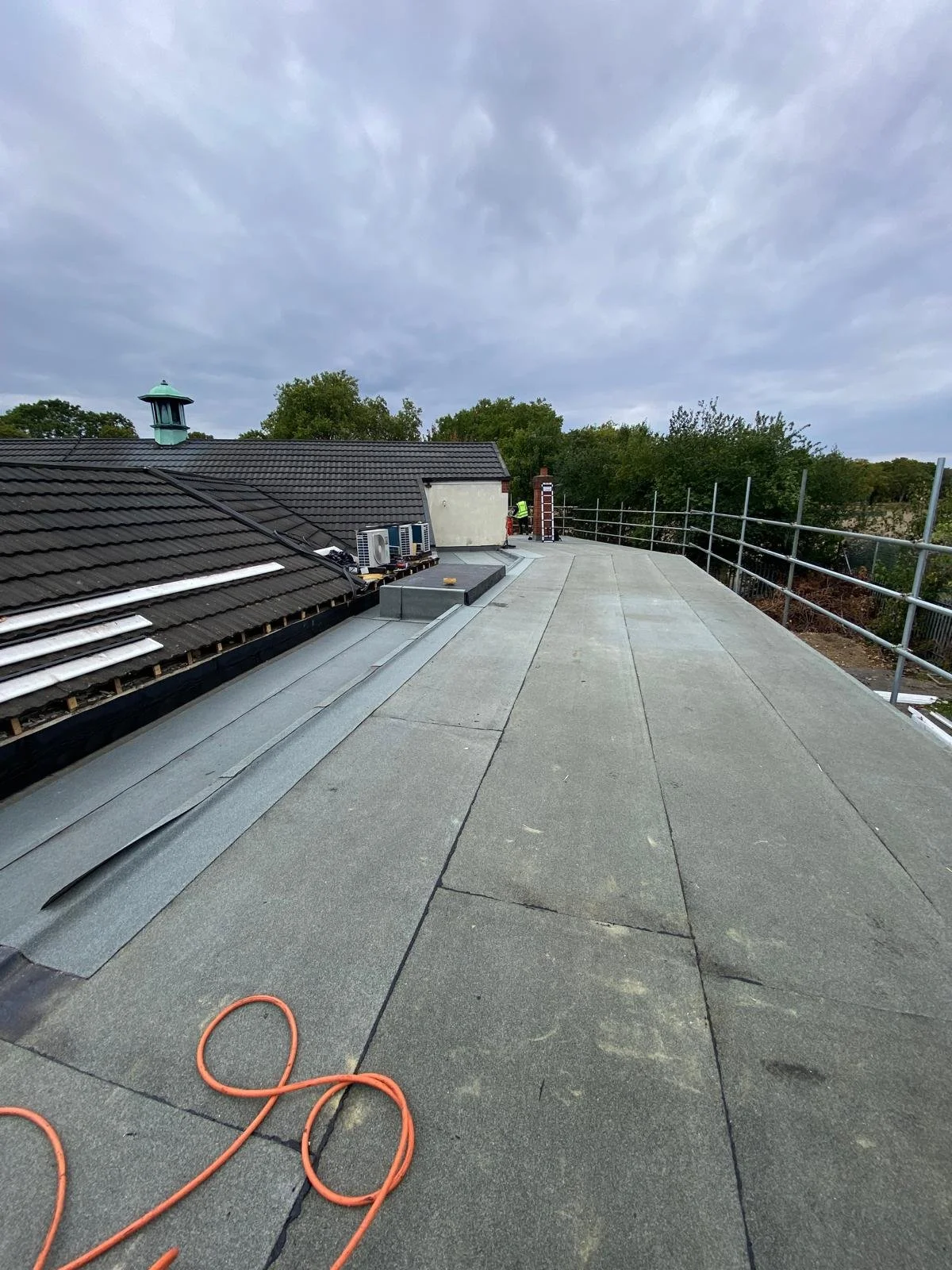 Roof construction site with a flat rooftop, construction materials, orange extension cord, and a worker in the background during daytime.
