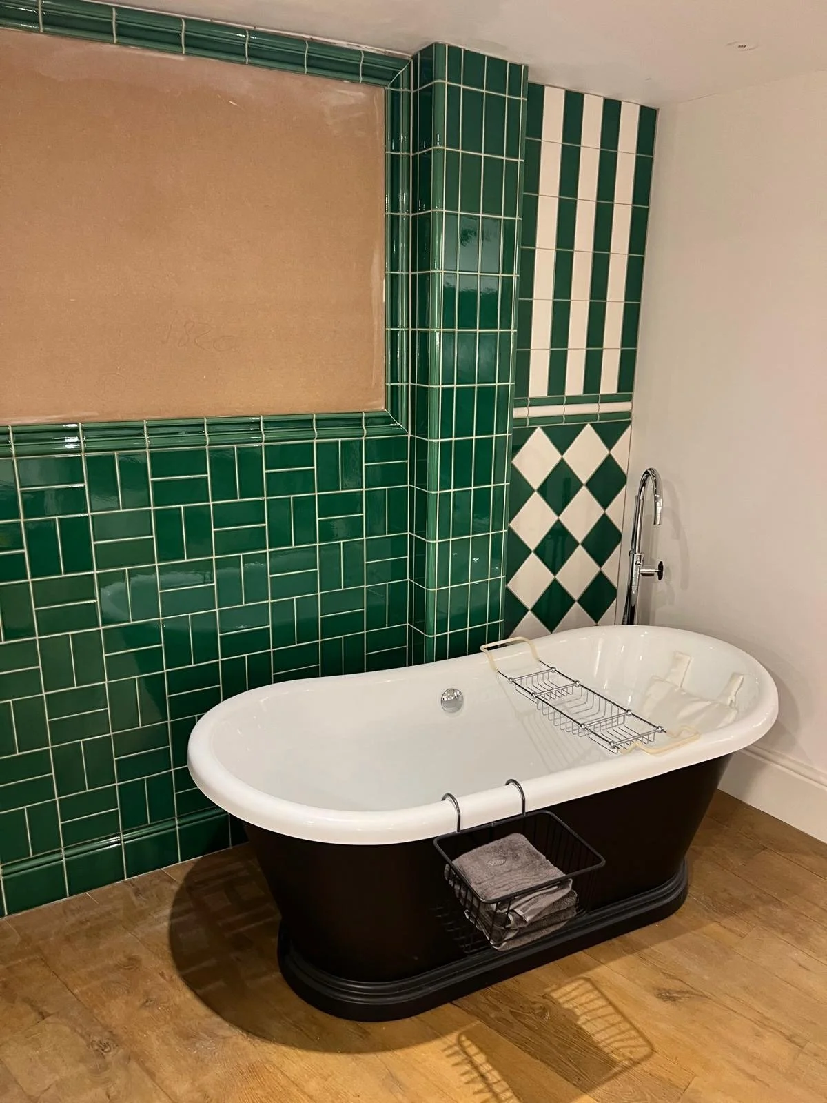 A bathroom with a black clawfoot bathtub, a wire bathtub rack, and a basket with towels underneath. The wall behind the tub is decorated with green and white tiles in geometric and striped patterns.