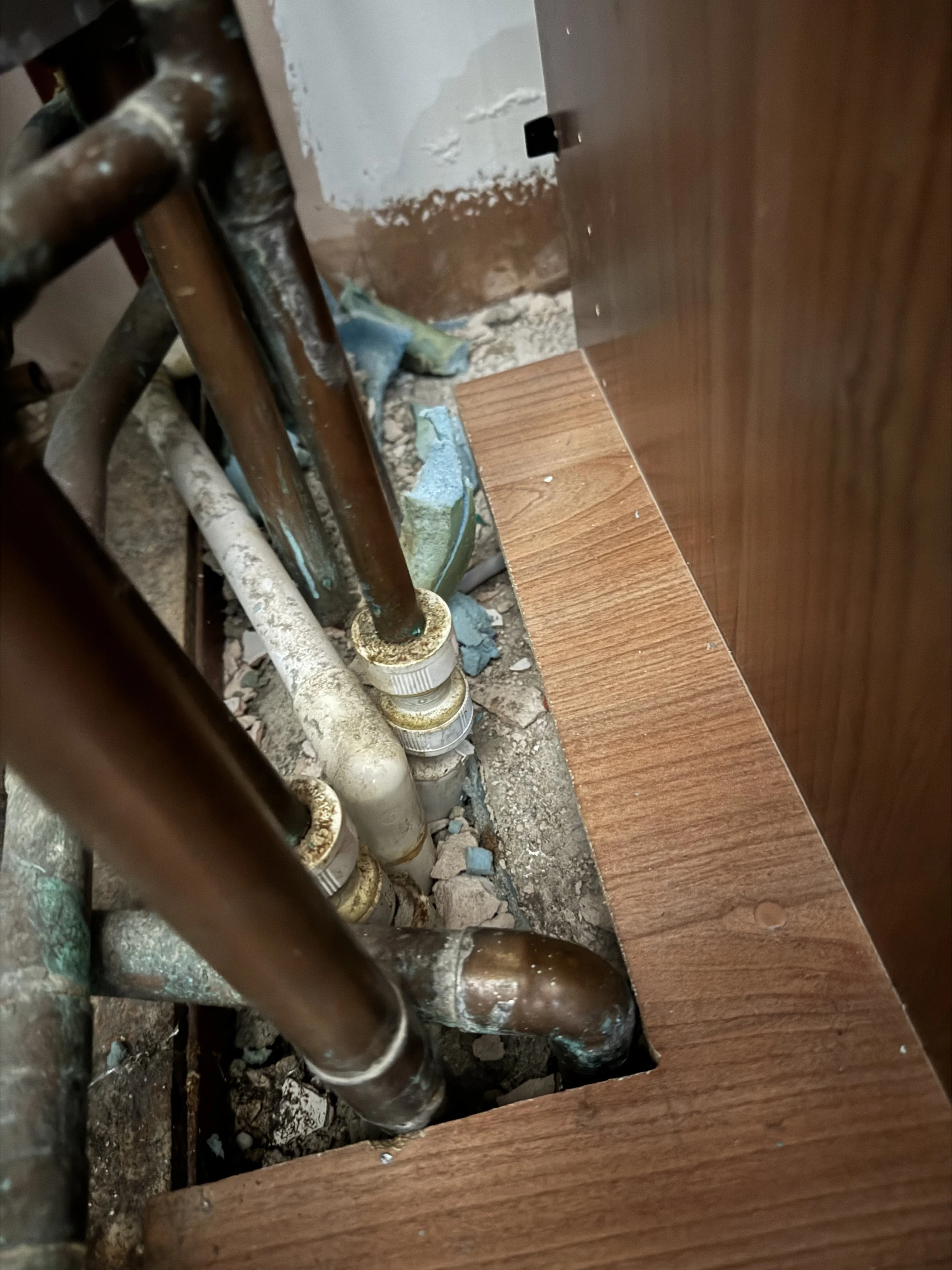 Close-up of plumbing pipes and fittings behind a wooden panel, with visible dirt and debris.
