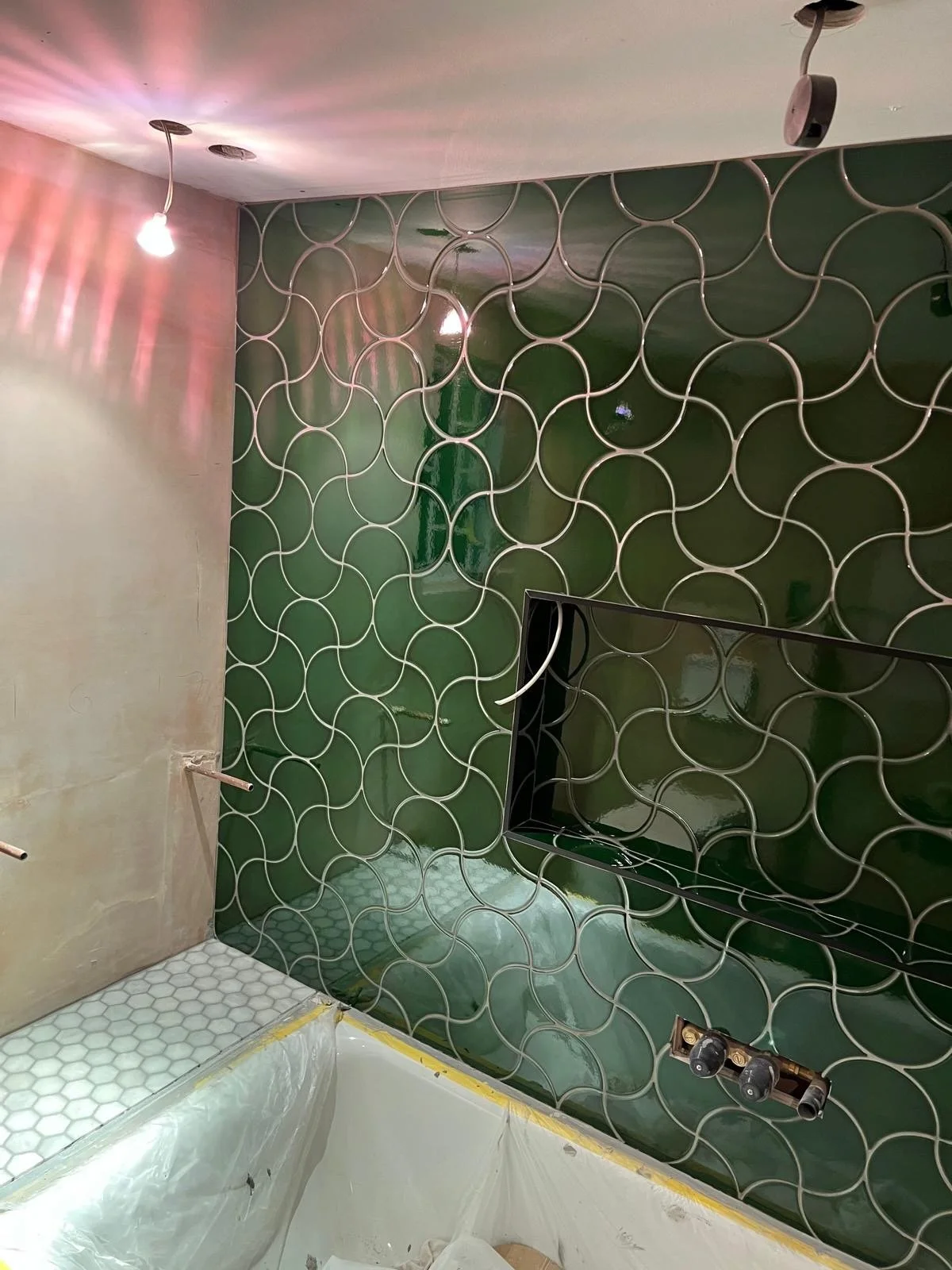 Interior of a bathroom under renovation featuring a wall with green decorative tiles and a rectangular cutout, a white hexagonal tile floor, and exposed plumbing fixtures.