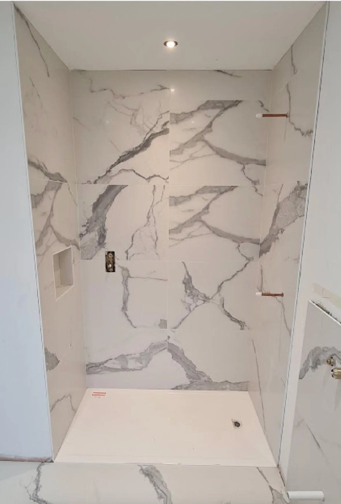 The image shows the interior of a shower with large white marble-like tiles with gray veining on the walls and floor. There is a small built-in niche for shower products, and wall hooks are installed on the right wall. The ceiling has a recessed ligh