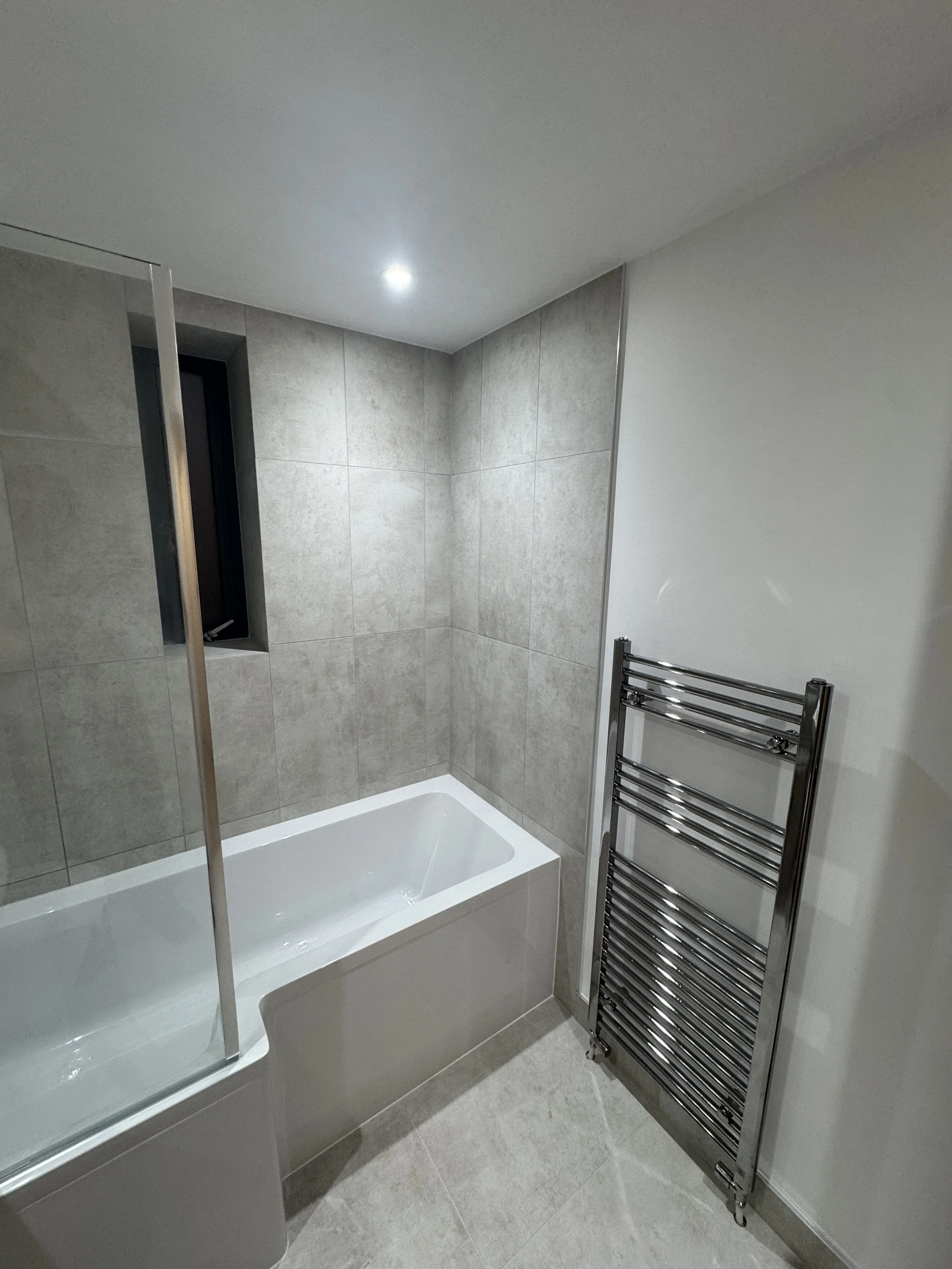 A modern bathroom with a bathtub, gray tiled walls, a small window, and a shiny silver heated towel rack.